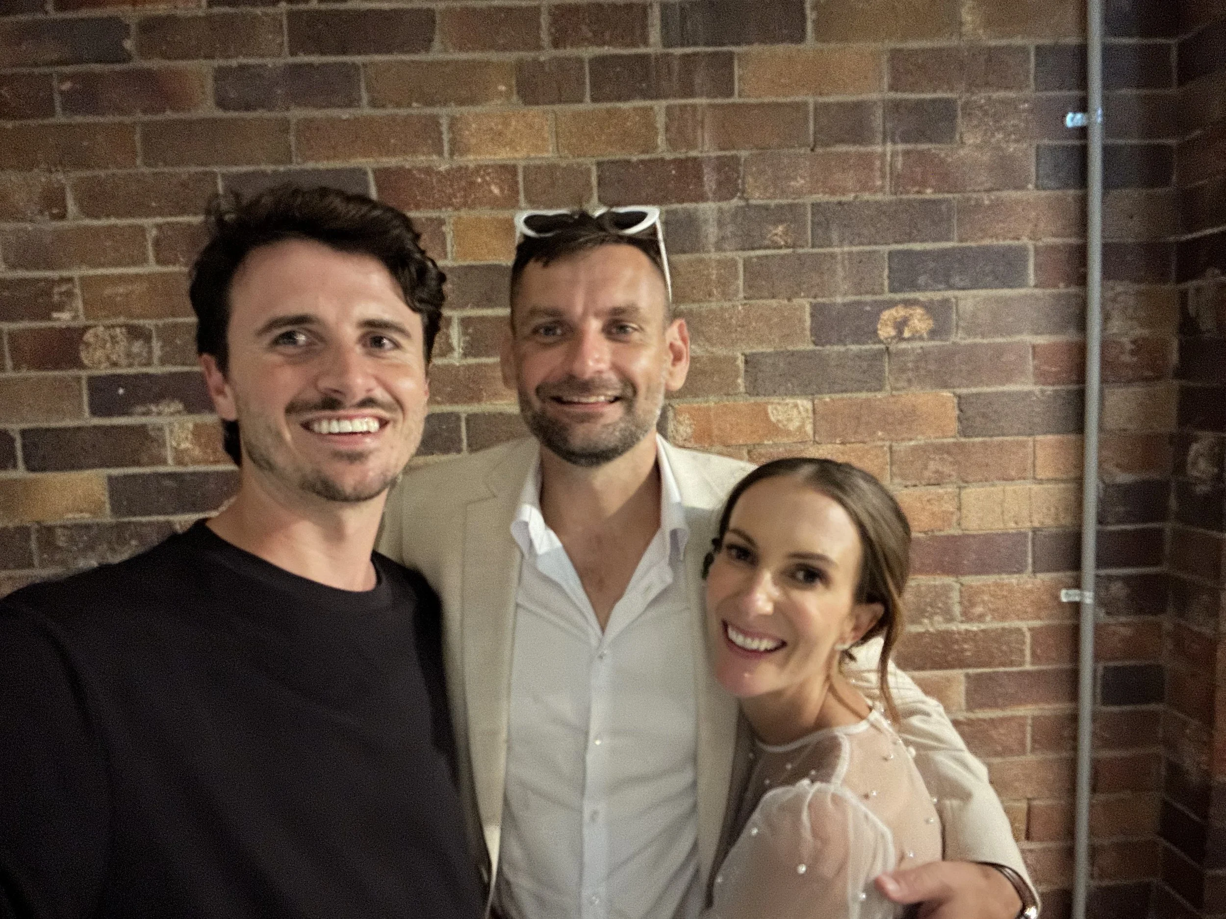 Three people posing for a photo in front of a brick wall. A man with dark hair and a black shirt is on the left, a smiling man with sunglasses on his head and a white shirt in the middle, and a woman with red hair and a sheer light-colored blouse on the right.