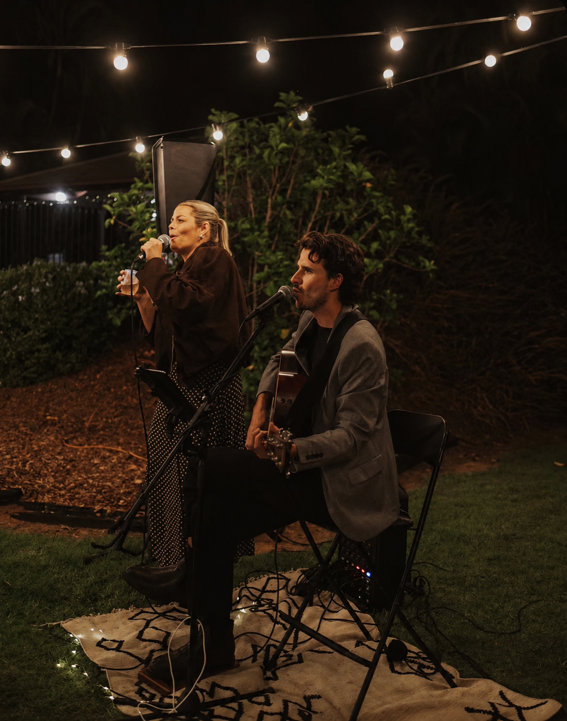 Two musicians performing outdoors at night under string lights, with one playing guitar and singing, and the other singing into a microphone with a drink in hand.