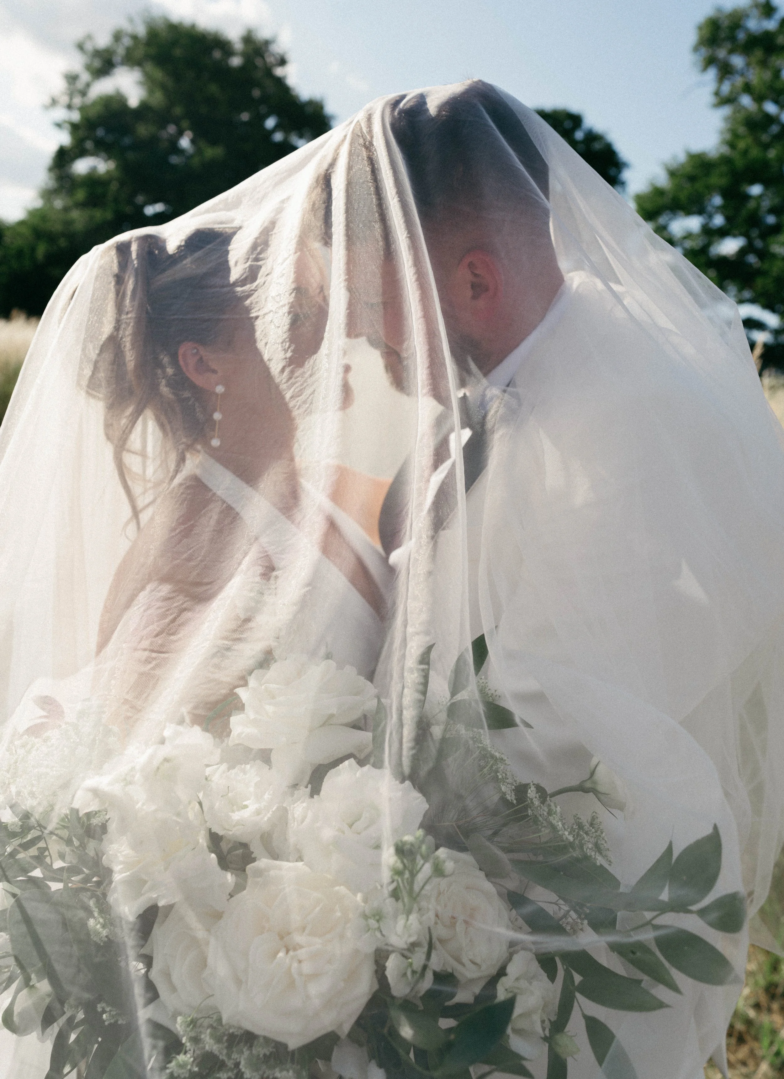 A bride and groom face each other beneath a wedding veil, holding a bouquet of white flowers outdoors with trees in the background.