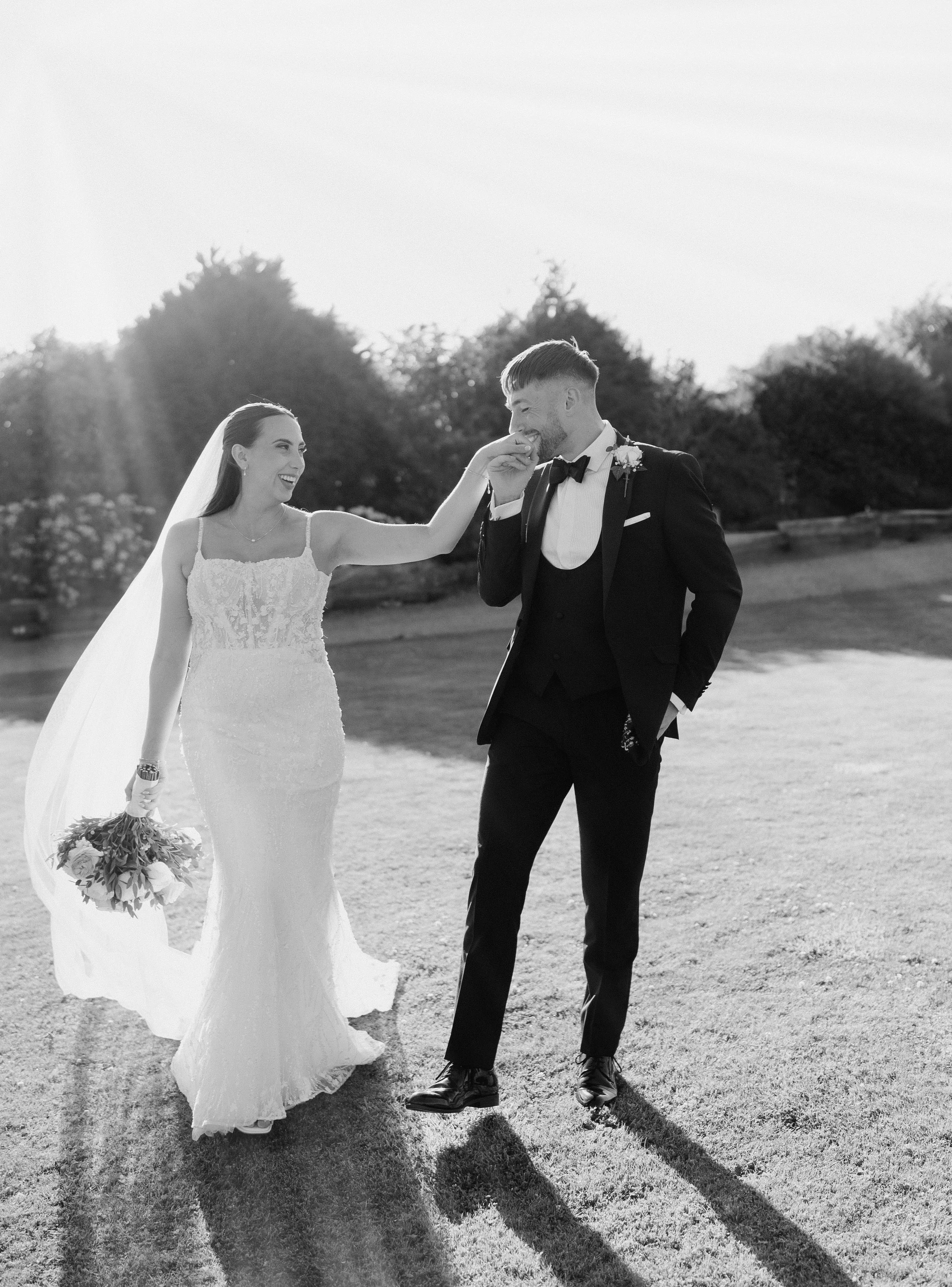 A bride and groom sharing a joyful moment outdoors, with the bride playfully touching the groom's face as they smile at each other, in black and white.