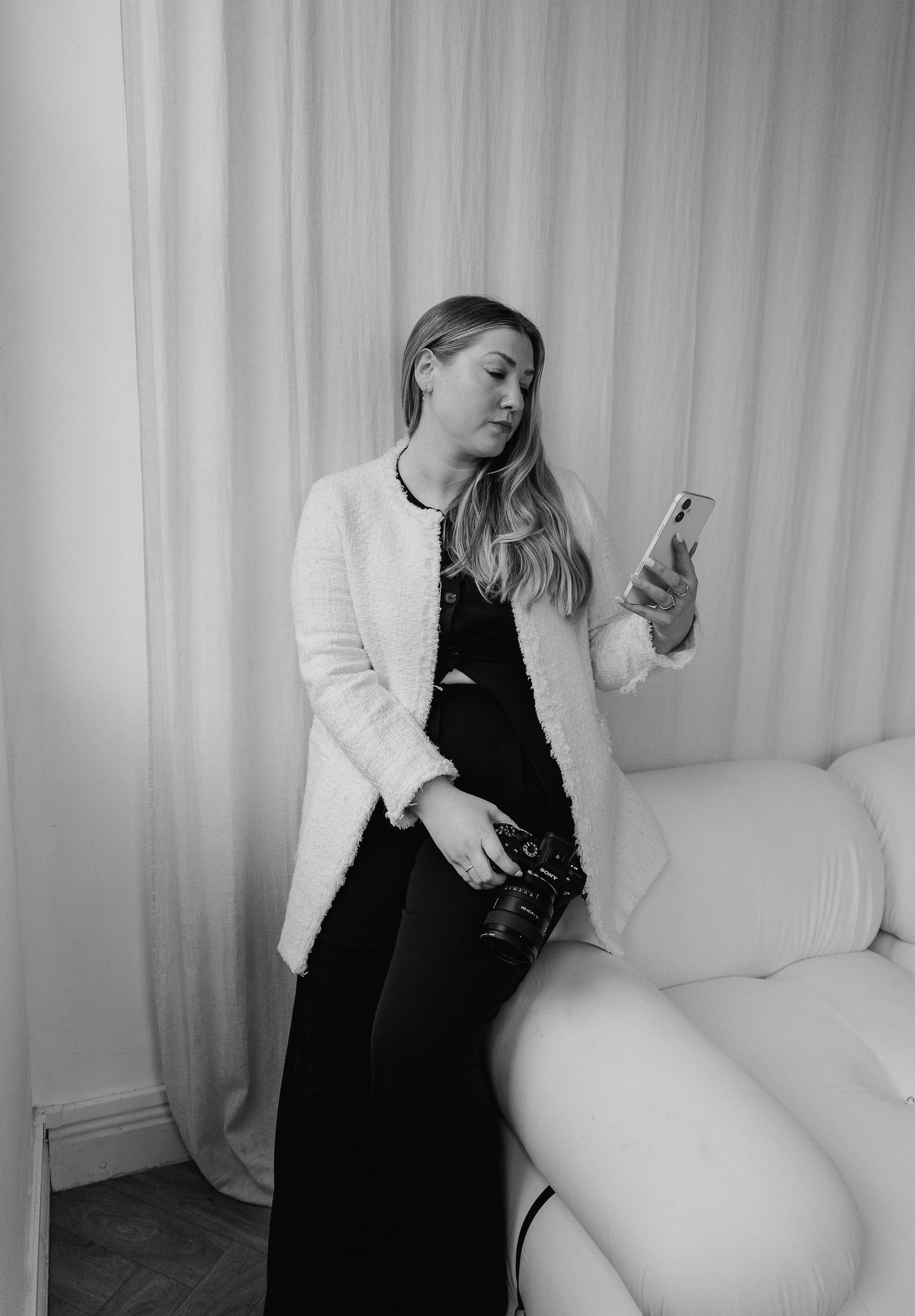 A woman with long hair, wearing a light-colored coat, standing inside, holding a camera in one hand and looking at her phone.