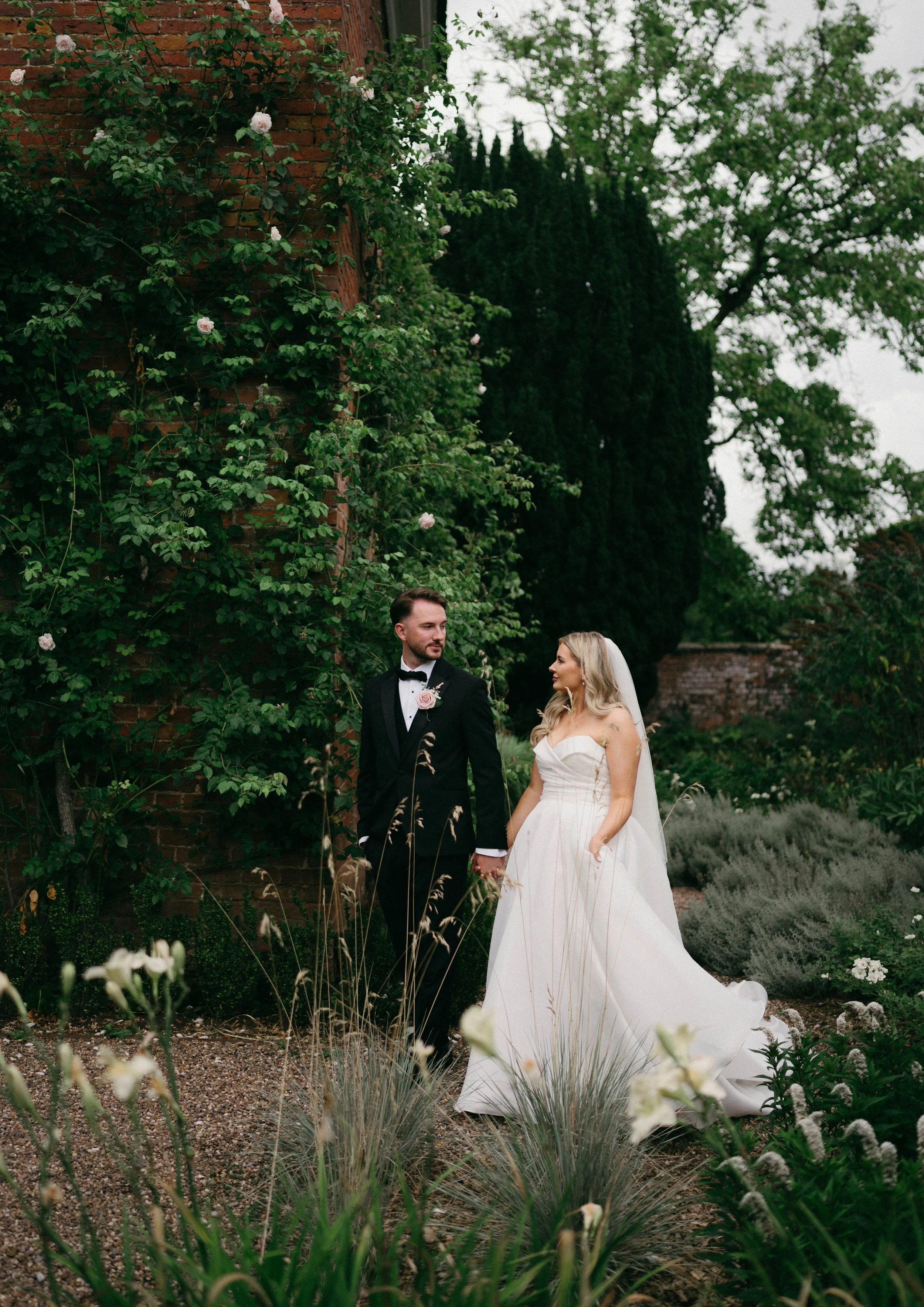 A bride and groom holding hands in a lush garden with greenery and flowers, the bride in a strapless white wedding gown with a veil, and the groom in a black tuxedo with a bow tie.