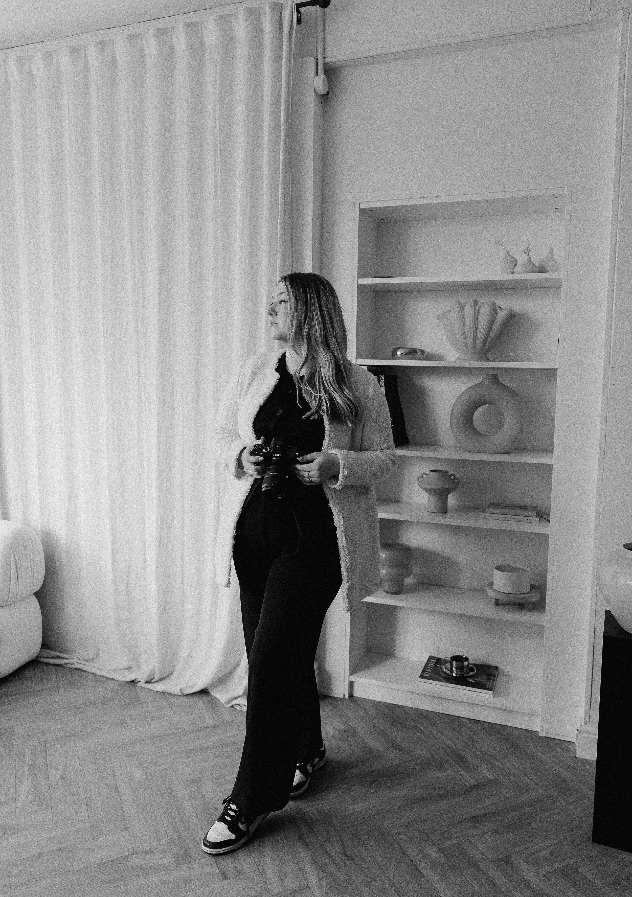 A woman standing indoors holding a camera, with a shelf filled with decorative ceramics and books behind her, and a curtain to her left.