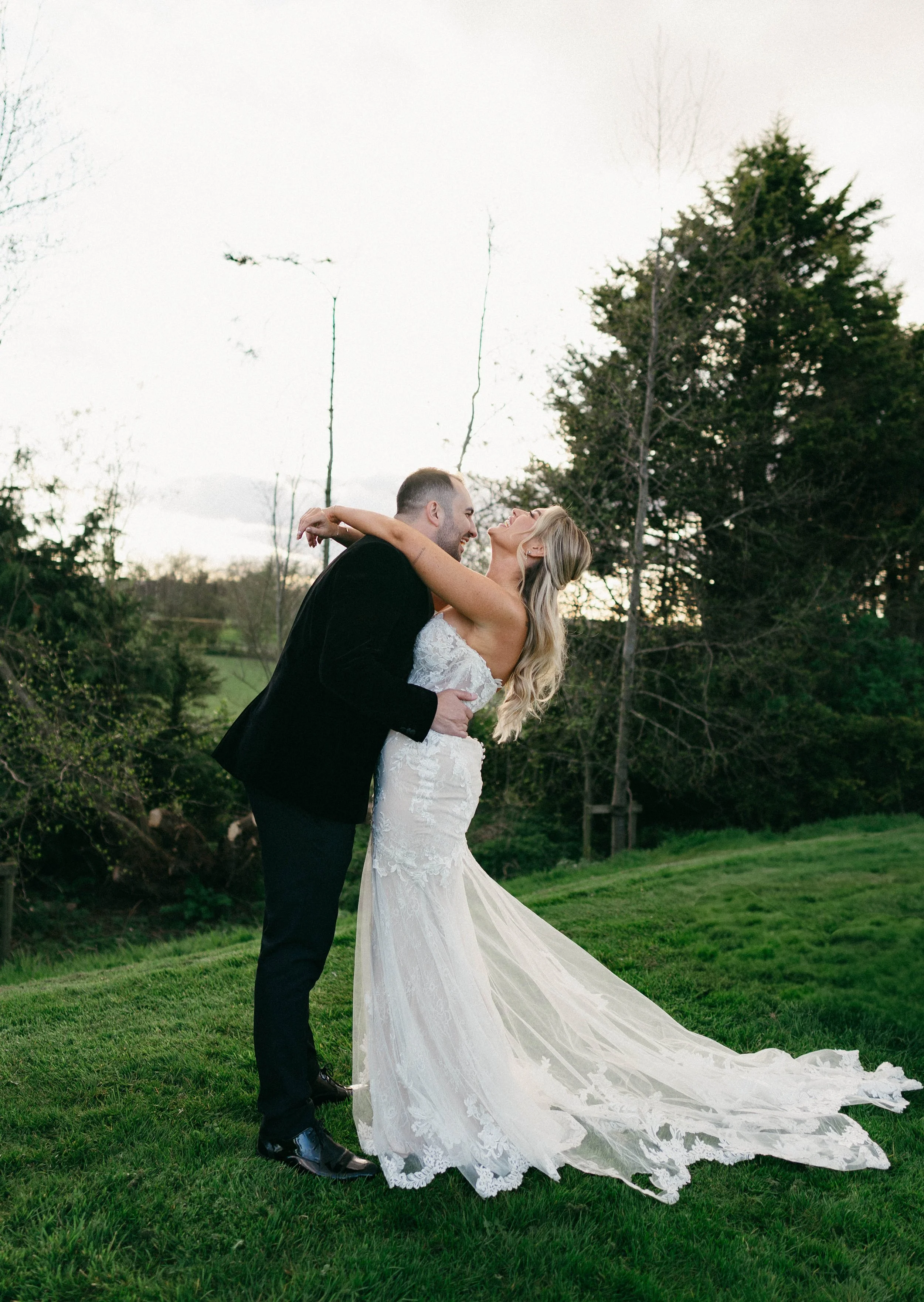 A bride and groom dancing outdoors on grass, smiling and looking at each other, with trees and a cloudy sky in the background.