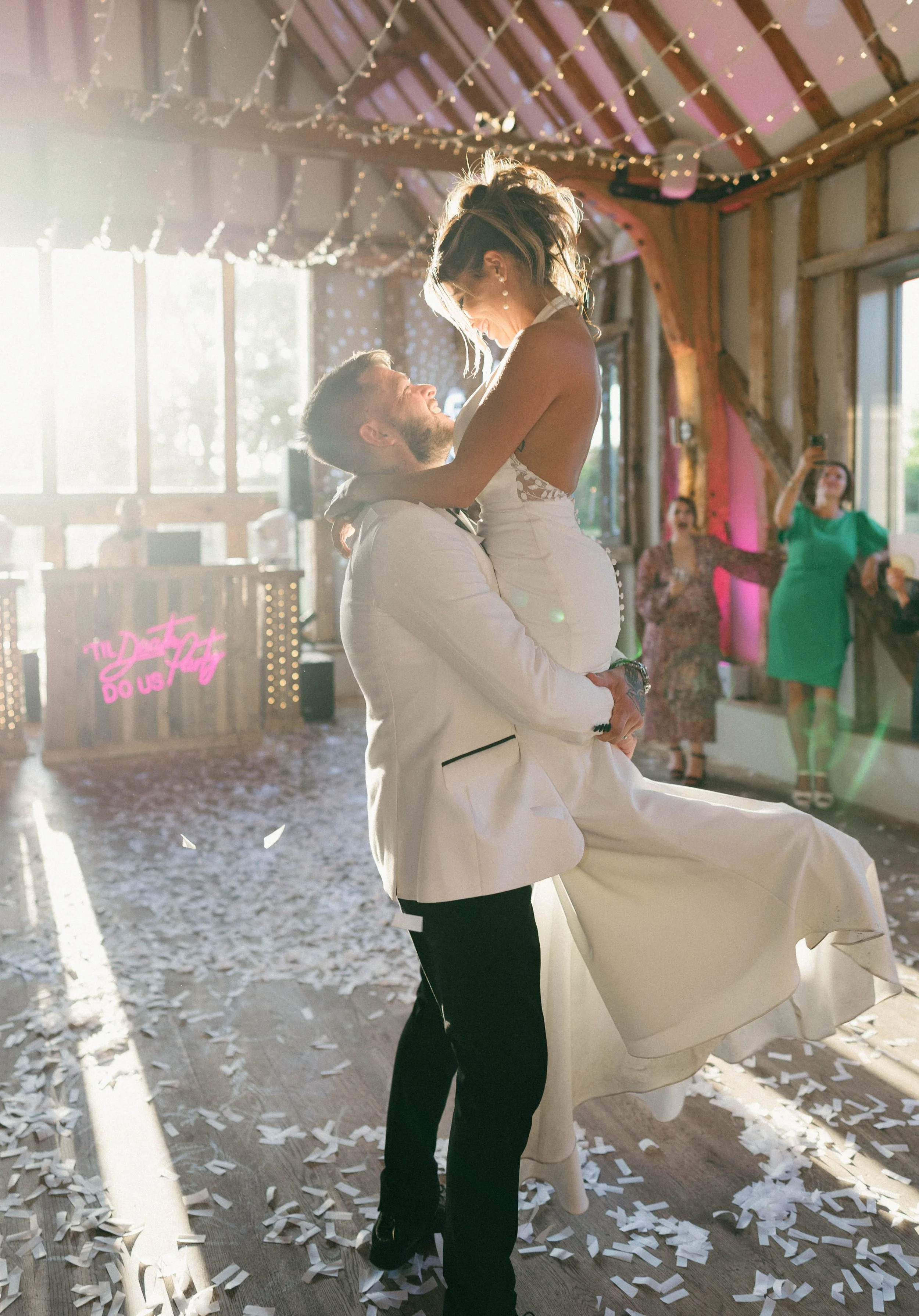 A bride and groom dance during their wedding reception, with the groom holding the bride up in an indoor barn setting decorated with string lights and a neon sign reading 'The Doot Do Us Part.'