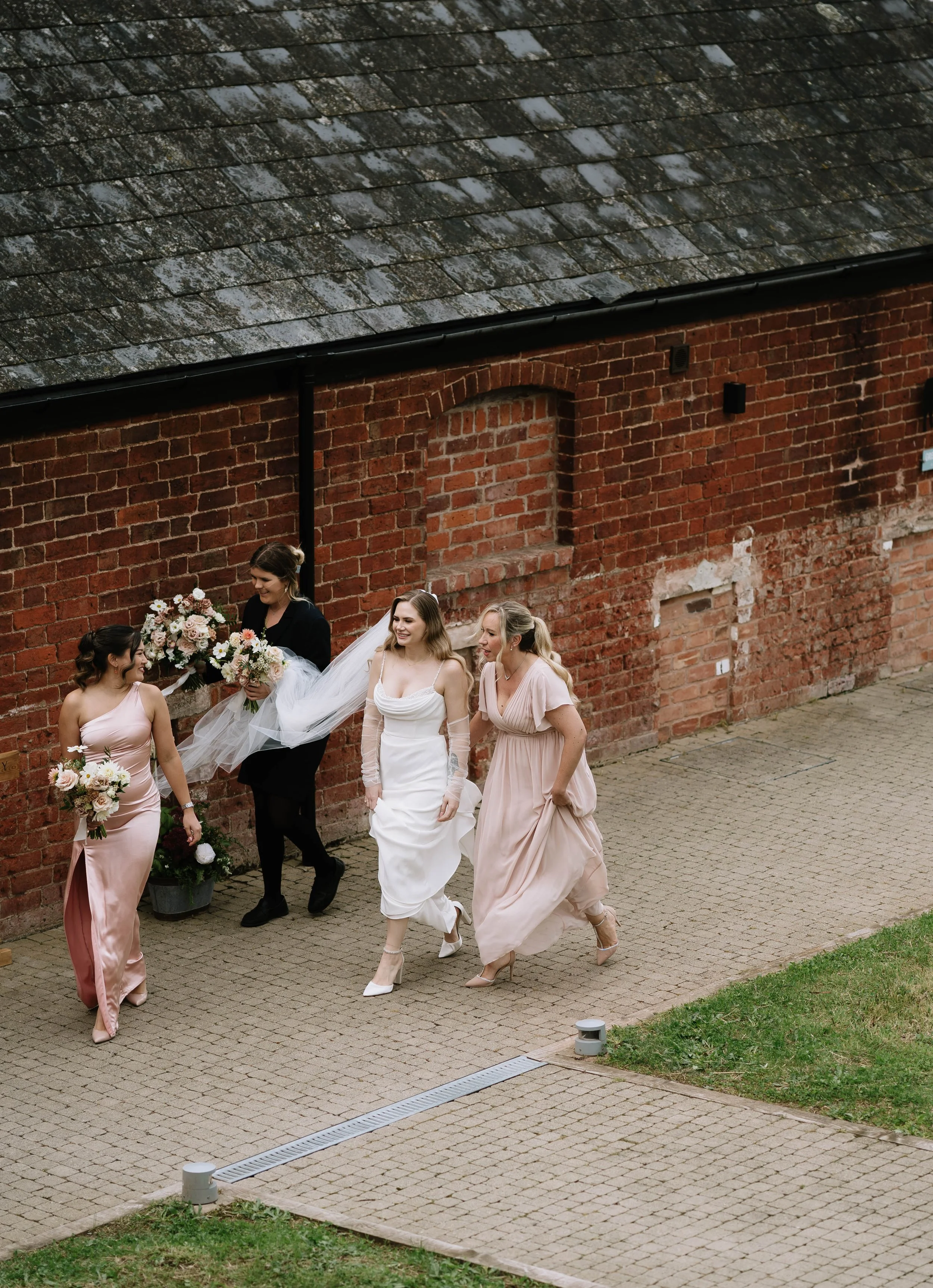 A bride and her bridesmaids walking outside near a red brick building, with one bridesmaid holding two bouquets of flowers, smiling and engaging in conversation.
