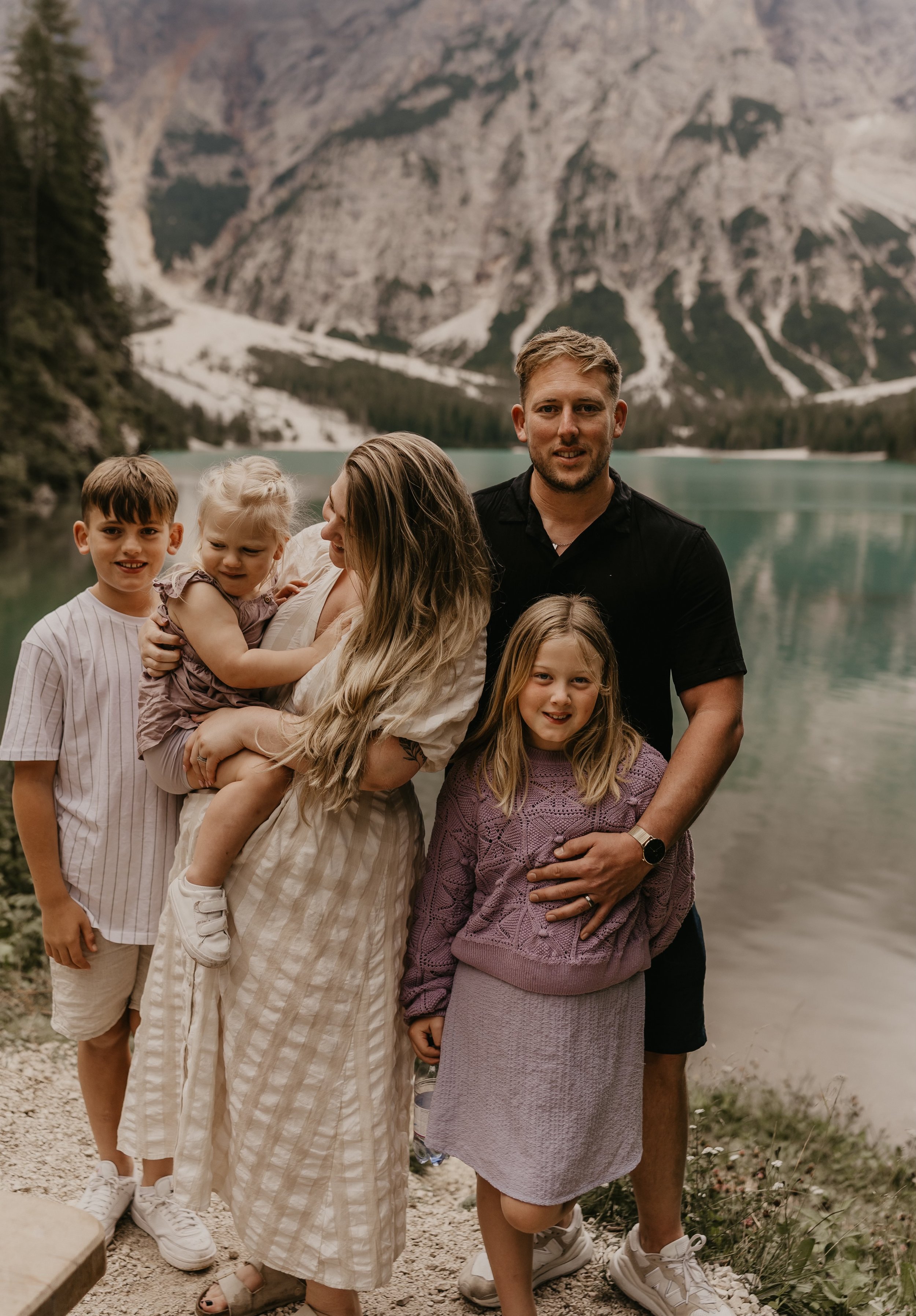 A family of five posing by a lake with mountains in the background. The father is standing on the right, holding a young girl in a purple sweater. The mother is in the middle, holding a young girl in a purple dress. A boy in a striped shirt stands on the far left.