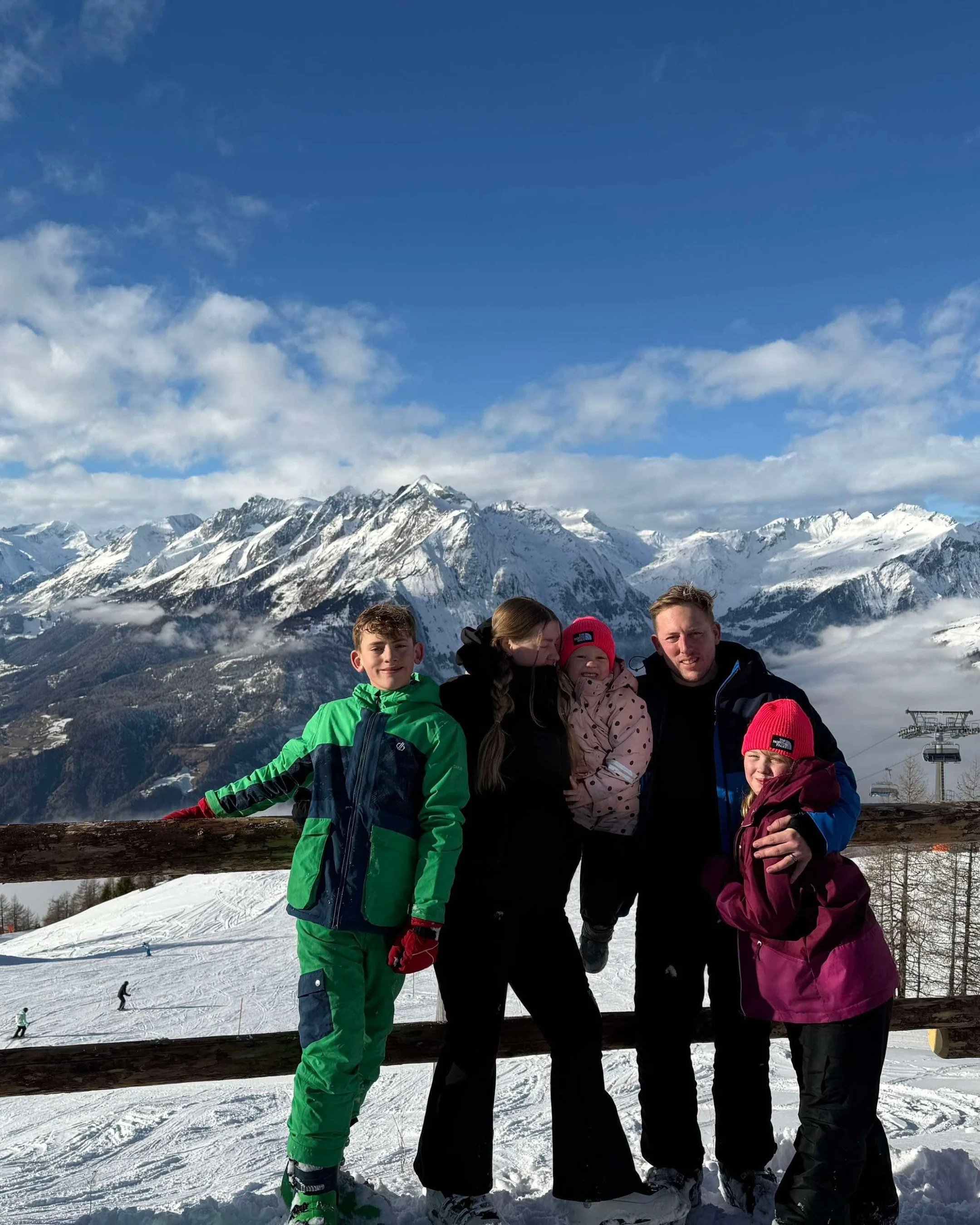 Family of five enjoying a snowy mountain view, with snow-capped peaks and ski lift in the background.