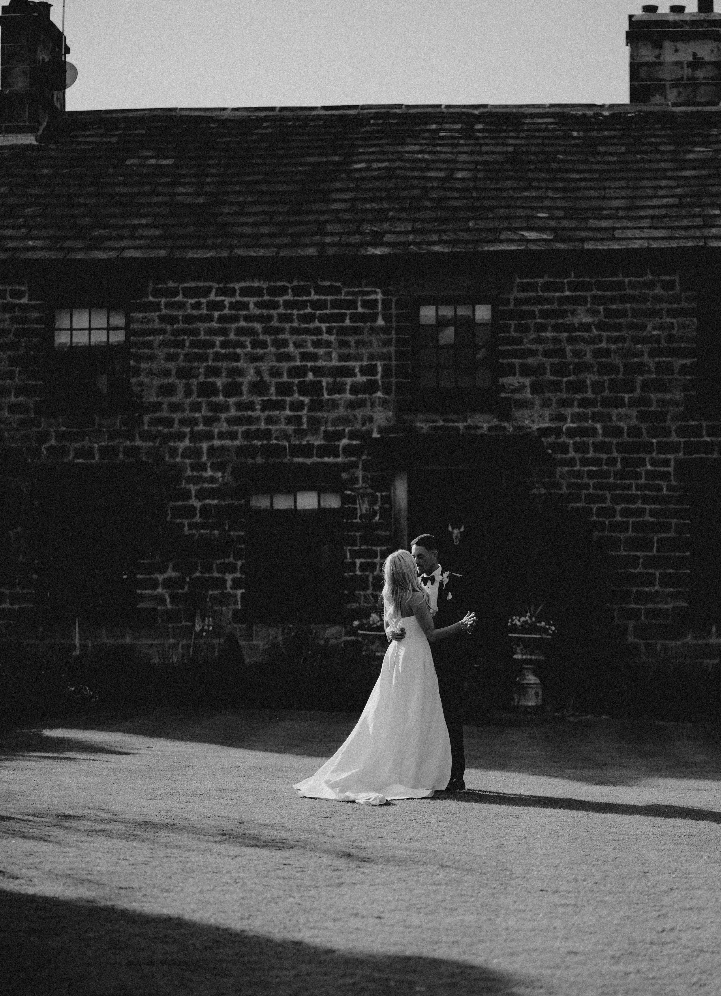 A black and white photograph of a bride and groom dancing outdoors in front of a brick building with a tiled roof.