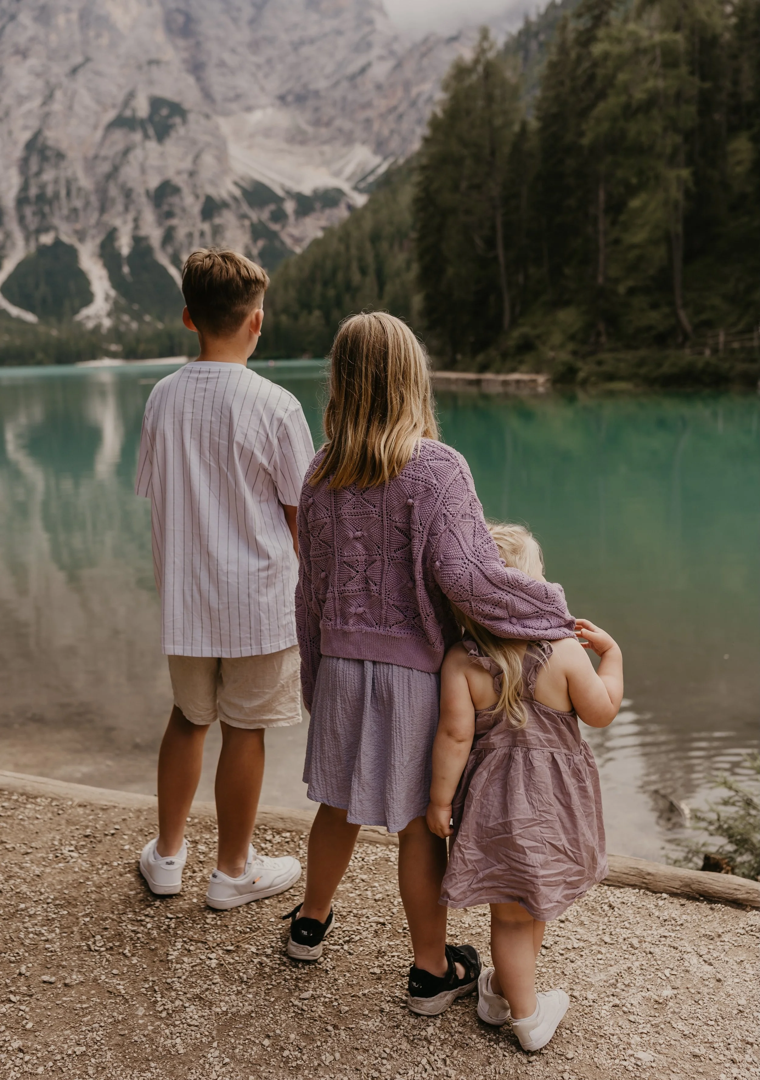 Three children, two girls and a boy, standing on a gravel shore by a lake, with mountains and trees in the background, looking at the water.