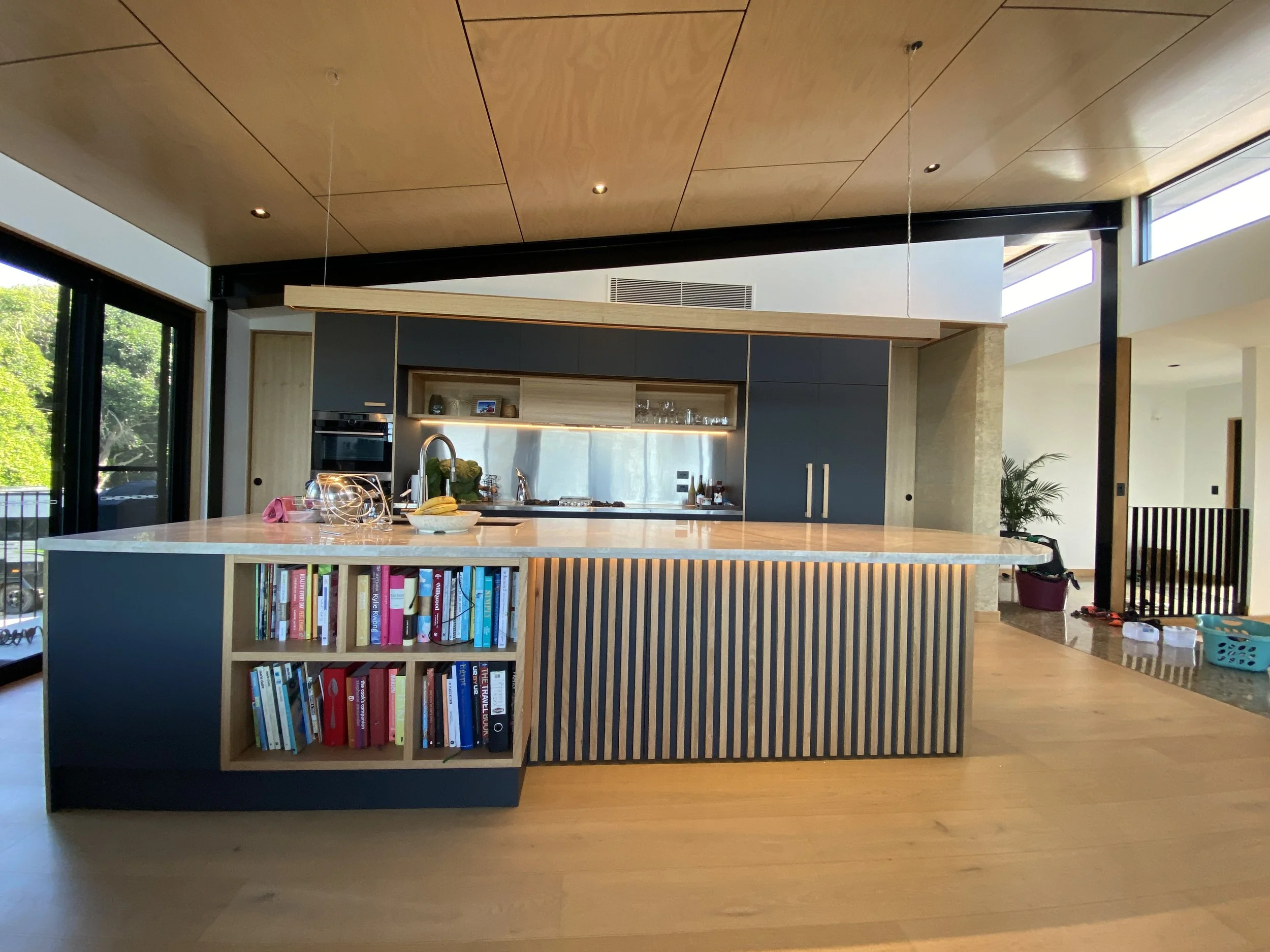 Modern kitchen with a large island, open shelves with books, and built-in appliances, in a bright space with wooden accents and sliding glass doors to the outside.