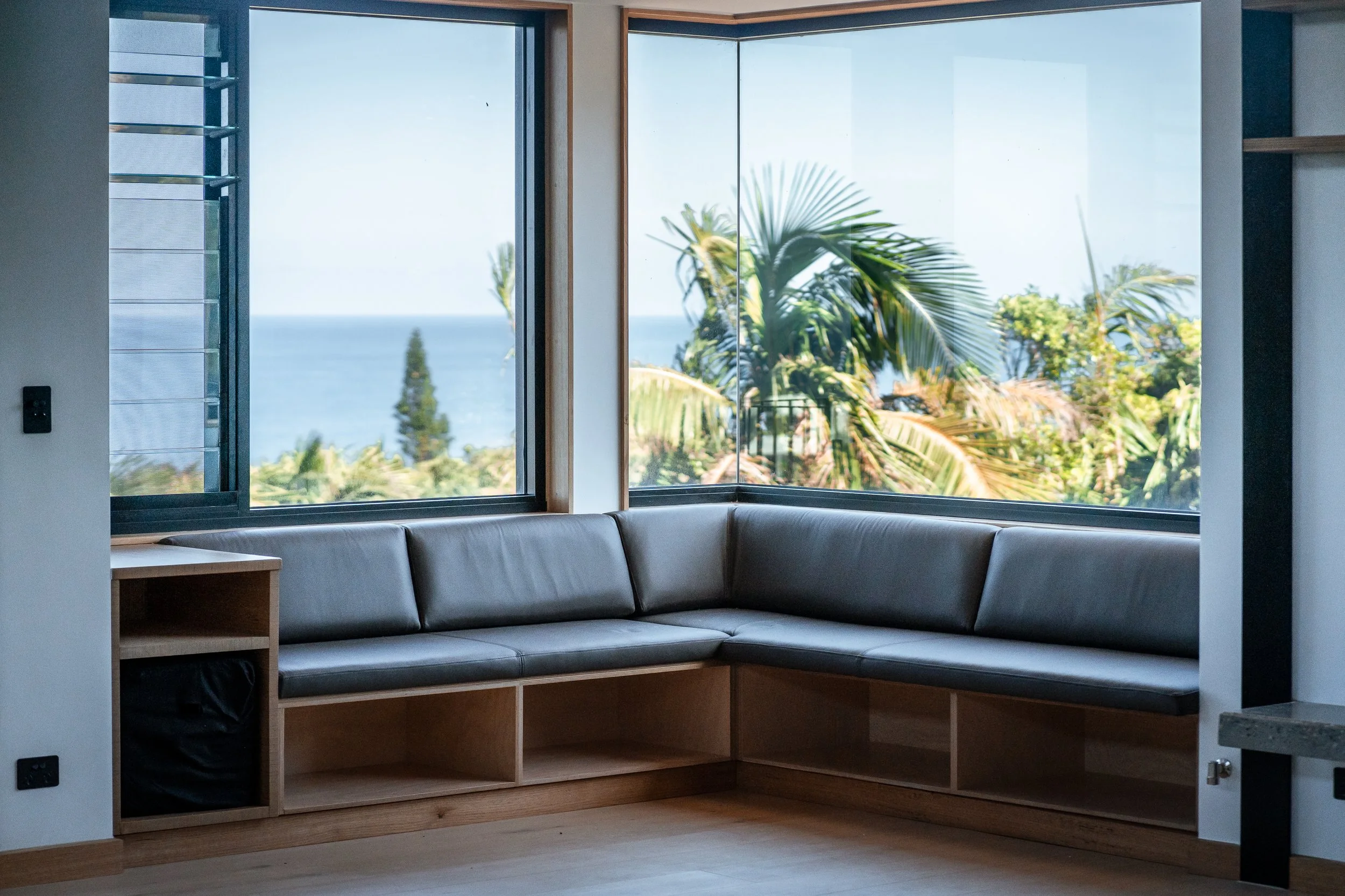 Interior view of a room with large windows showing a view of palm trees, the ocean, and a partly cloudy sky, with a modern black leather corner sofa and wooden shelving.