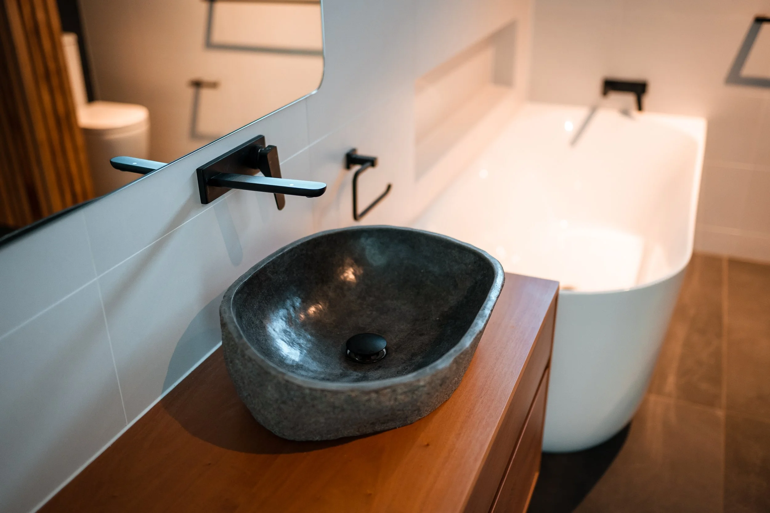 Modern bathroom with a black stone sink, wooden countertop, and a white bathtub in the background.