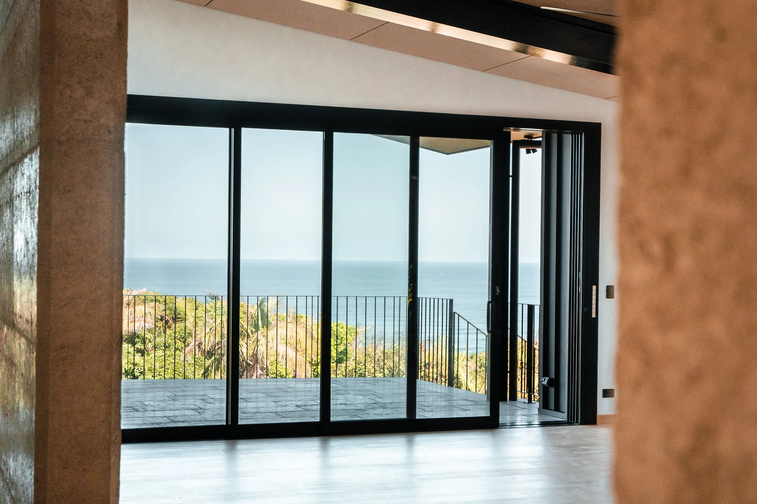 Interior of a modern room with large glass sliding doors opening to a balcony with a view of the ocean and trees.