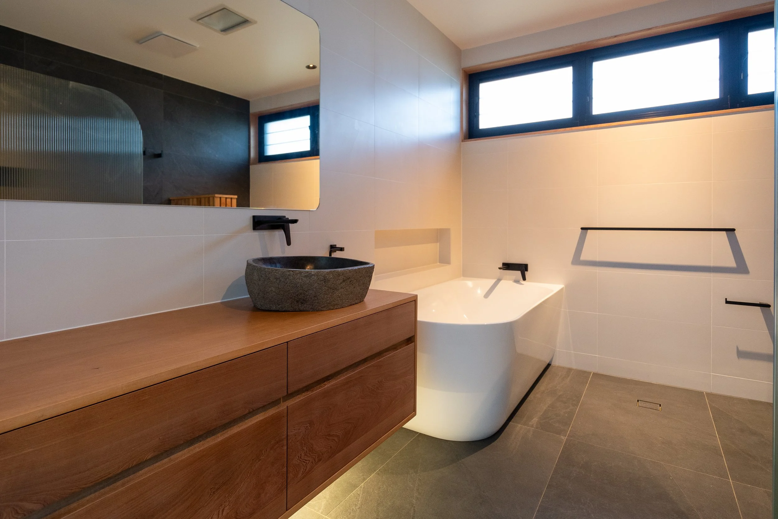 Modern bathroom with a freestanding bathtub, wooden vanity with black vessel sink, large mirror, and horizontal windows.