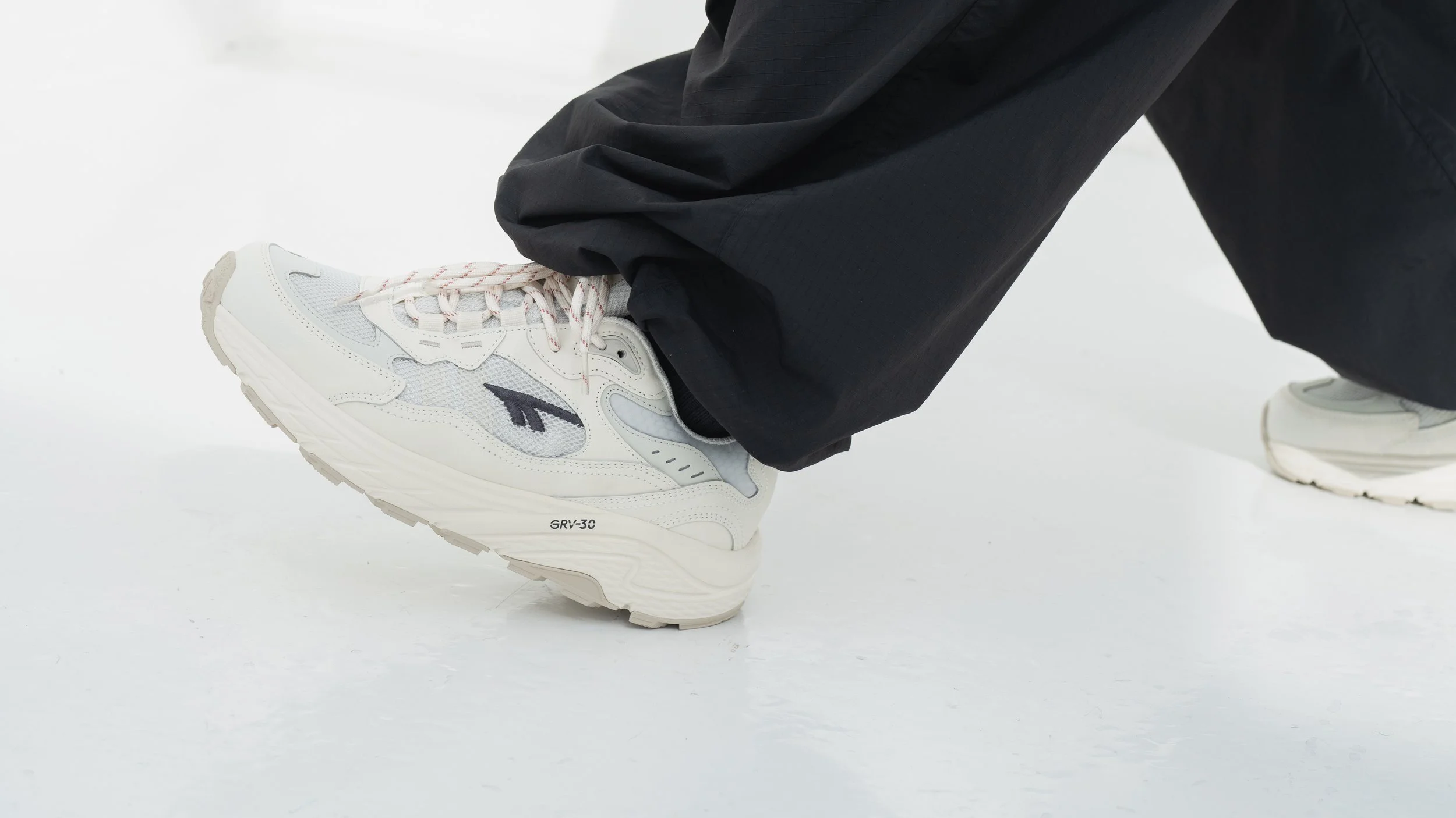 Close-up van een wit sportieve schoen met een navyblauw logo, gedragen door iemand in zwarte broeken op een witte vloer.