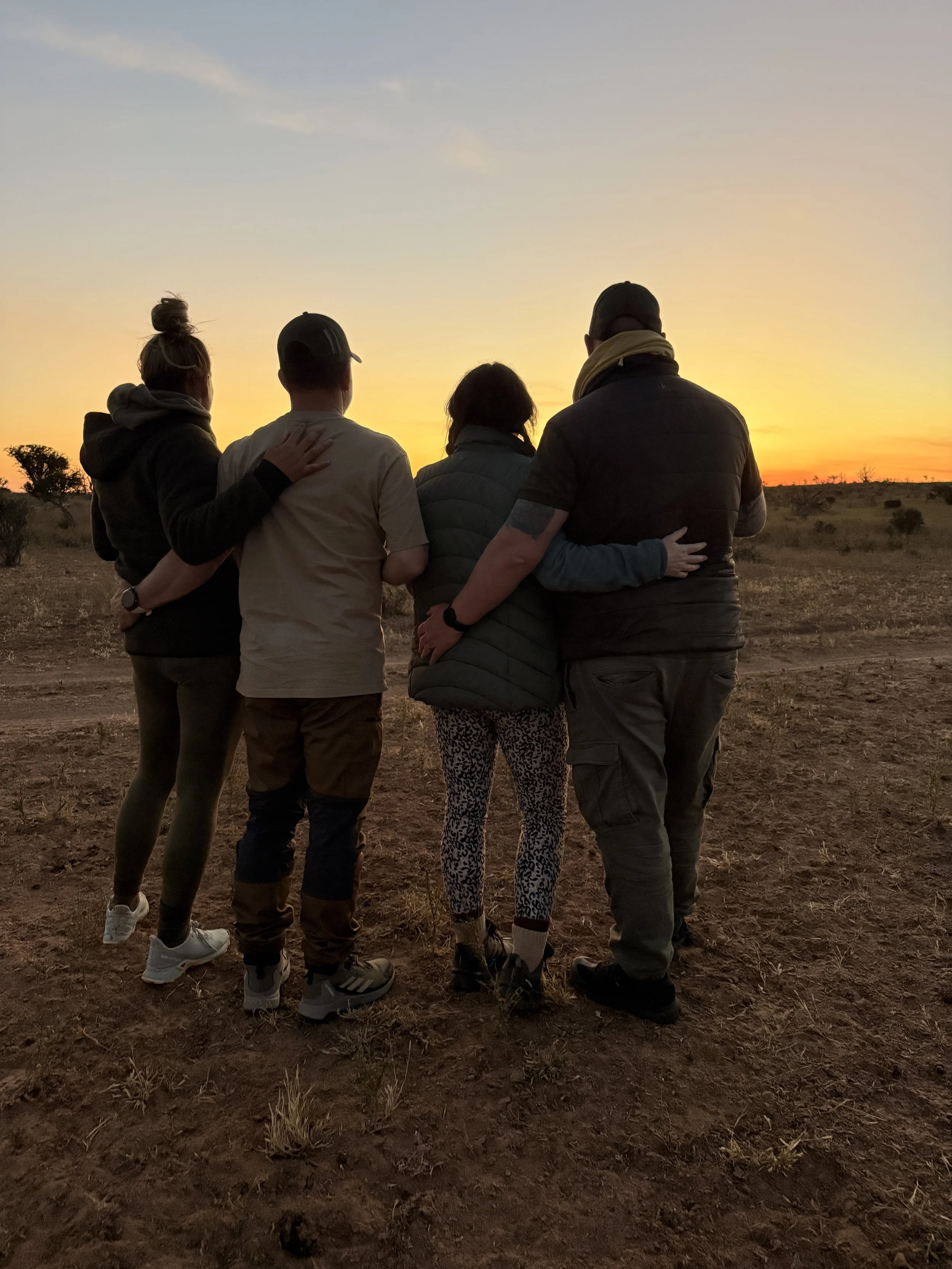 Group of five people standing closely together outdoors at sunset, facing away from the camera, in a barren landscape with sparse vegetation.