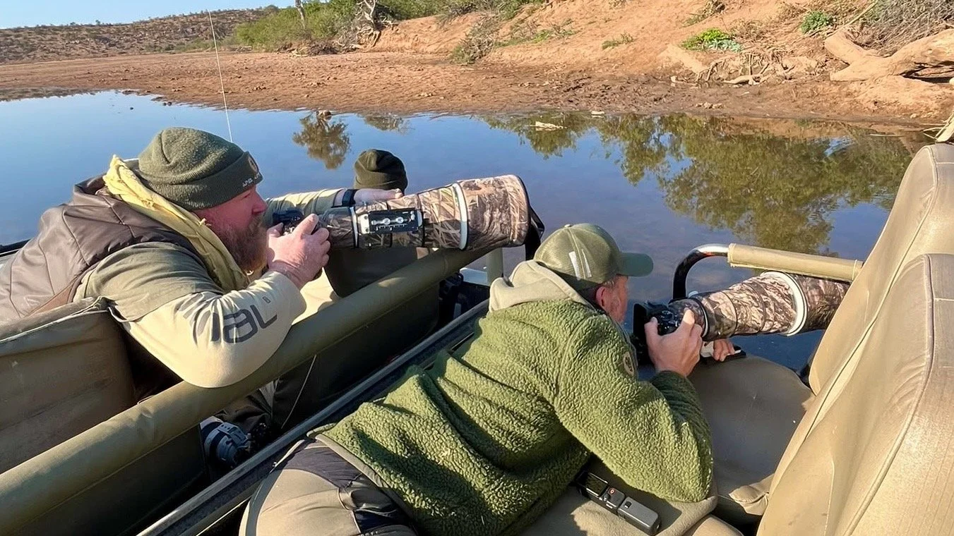Two men in an open boat, aiming rifles with scoped attachments, by a riverbank, possibly hunting or birdwatching.