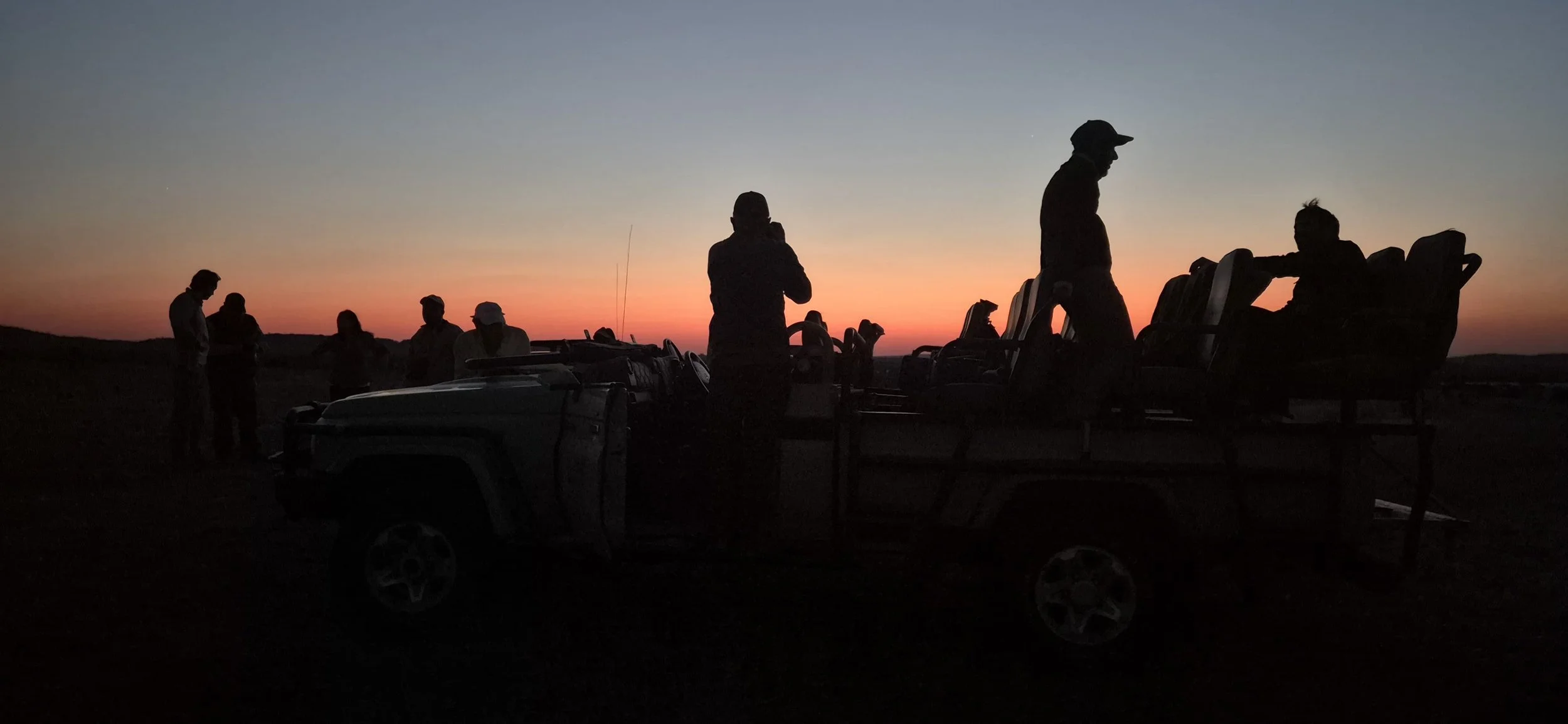 Silhouettes of people standing and sitting on a vehicle at sunset or dusk, with a colorful sky in the background.