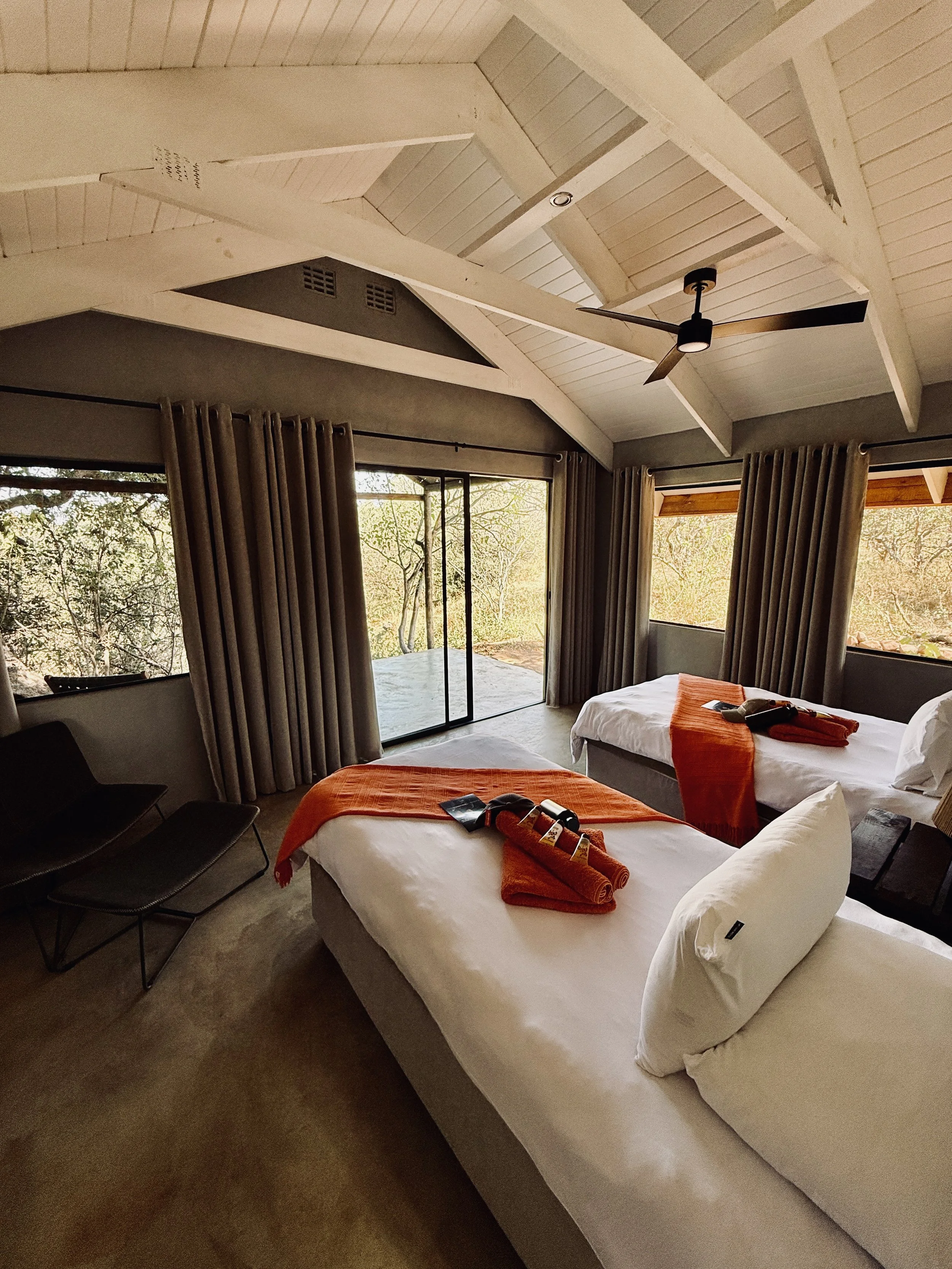A hotel room with two beds covered in white linens and orange throw blankets, with rolled orange towels and toiletries on top. The room has large windows with brown curtains, a sliding glass door leading outside, and a ceiling fan. Outside, trees and natural scenery are visible.