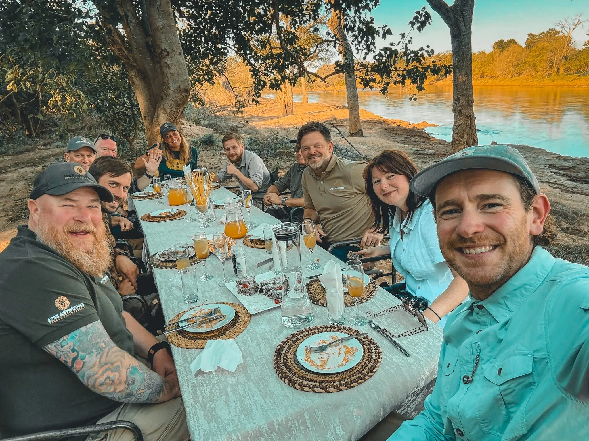 A group of eight people gathered around a long outdoor dining table by a river during sunset. They are smiling, with drinks and plates of food in front of them. The background features trees and a calm river, with the sky showing warm colors.