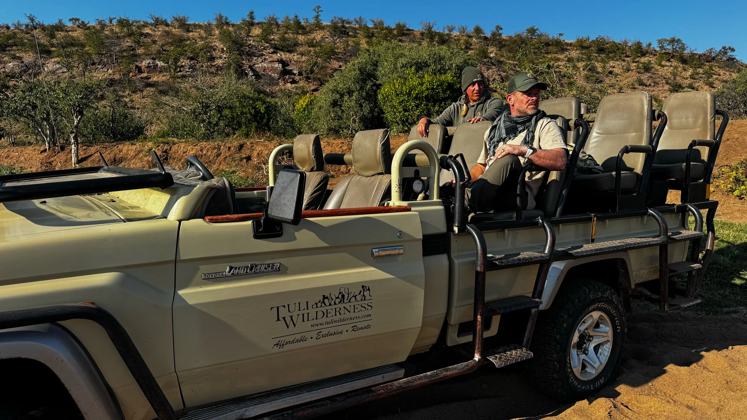 Two people sitting in an open safari vehicle in a wilderness area with dry hills and sparse trees in the background.