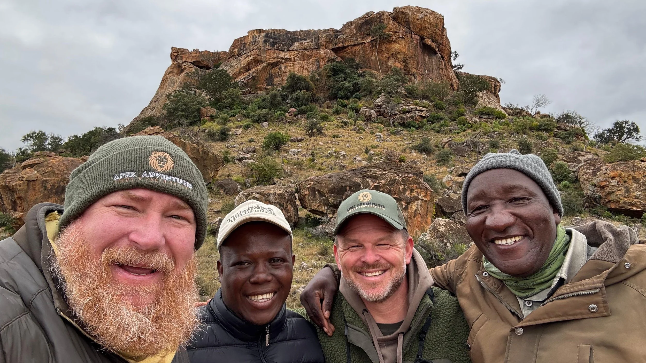 Group of five men outdoors taking a selfie with a rocky hill in the background, smiling and wearing outdoor gear.