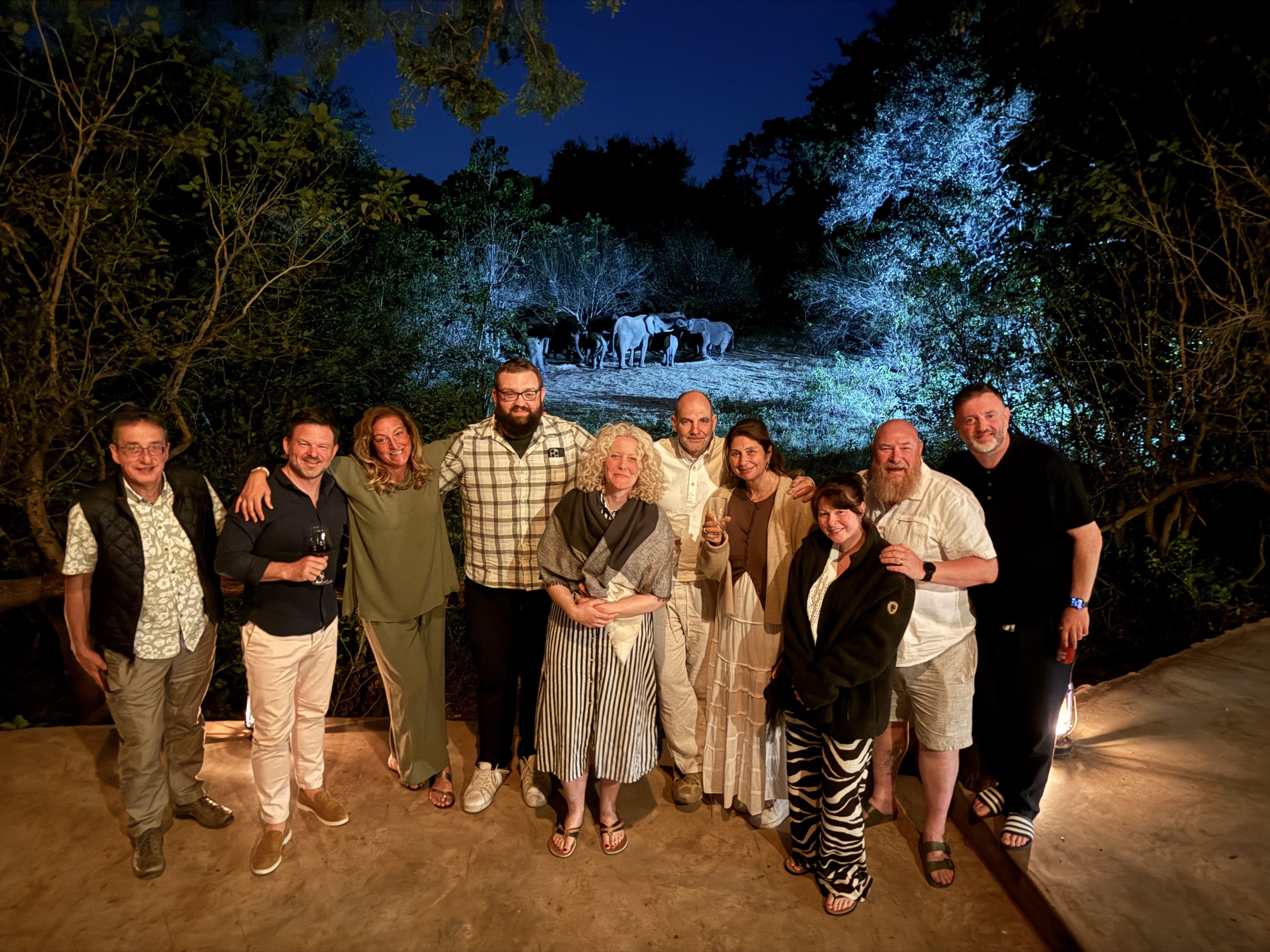 A group of people posing for a photo at night in front of a mural or projection of elephants in a forest scene with trees and a dark sky.