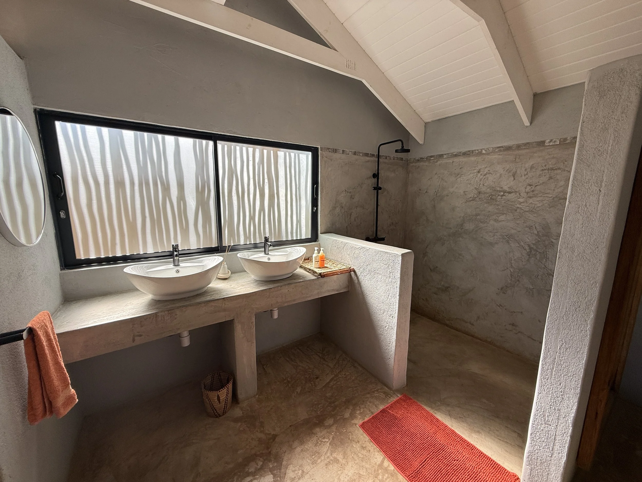 A bathroom with a large rectangular window, two white vessel sinks with faucets, a mirror, and a shower with a mounted showerhead. The bathroom features beige stone-like flooring and walls, a concrete countertop, and an orange towel hanging on the left.