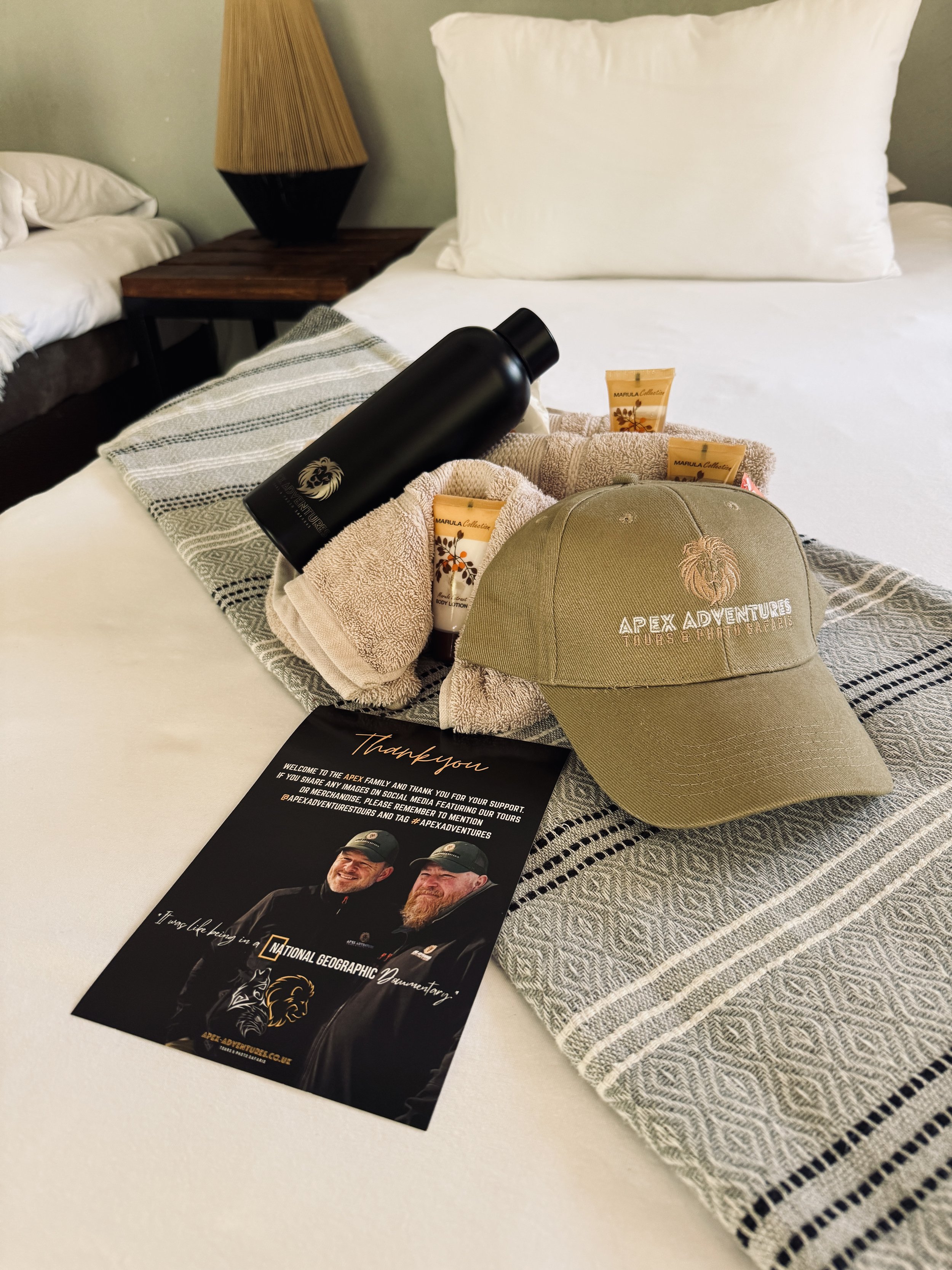 A hotel room bed with a white pillow, striped blanket, and a tray holding a black water bottle, skincare products, a beige cap embroidered with 'Apex Adventures,' and a thank you note from National Geographic Adventures. The room has a side table with a lamp and a second bed with white linens.
