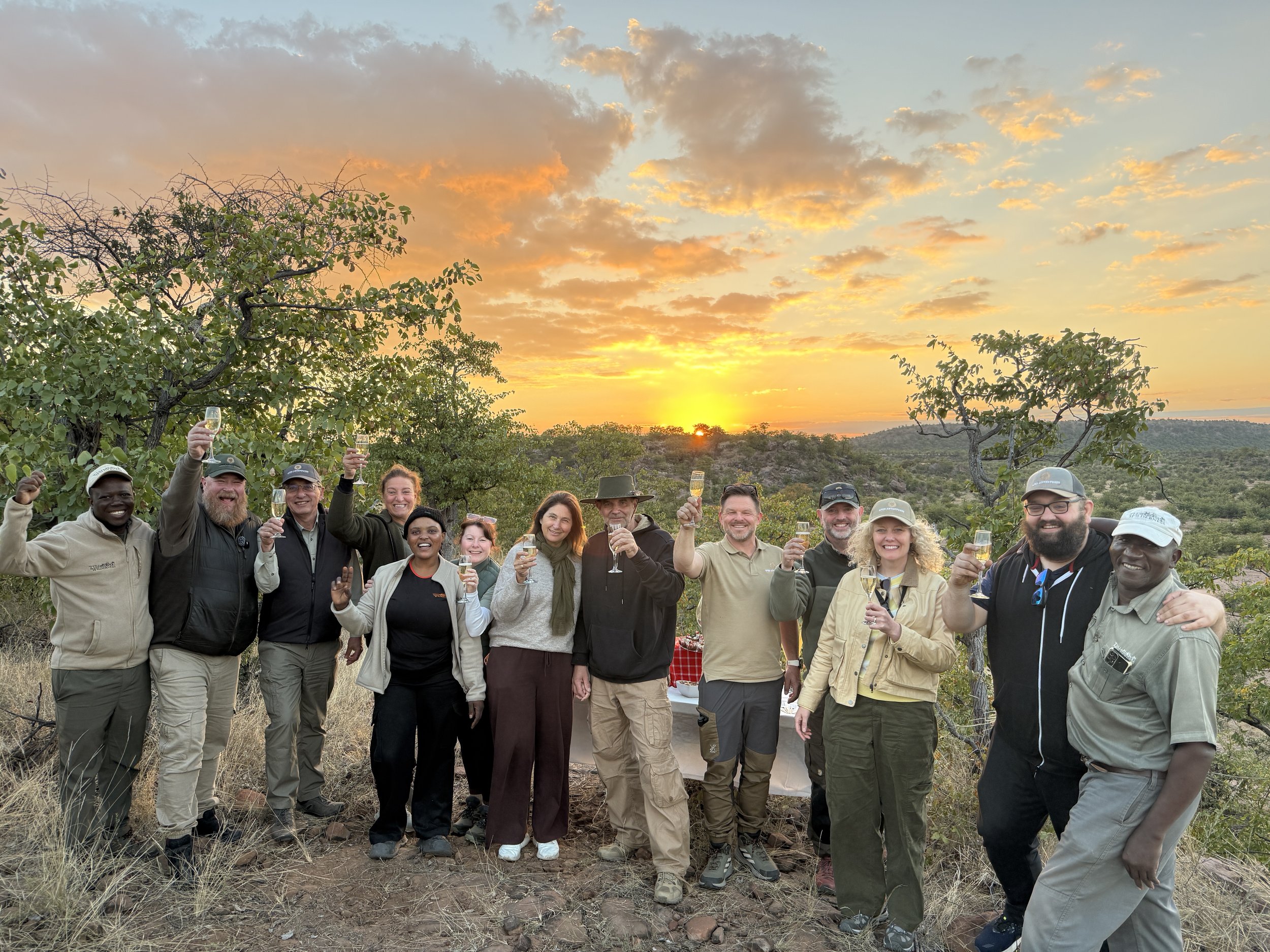 A group of people outdoors at sunset, celebrating with champagne in a savanna landscape with trees and hills.