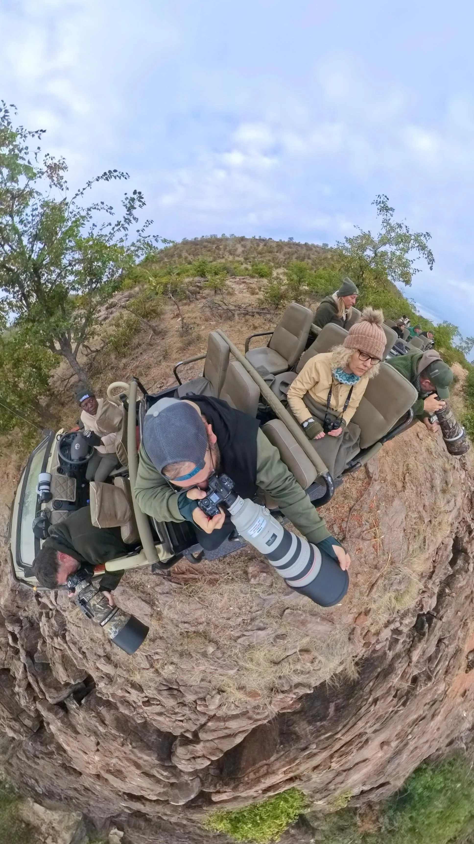 A group of people in safari vehicles observing and photographing wildlife in a rocky, canyon-like terrain with sparse vegetation and a partly cloudy sky.