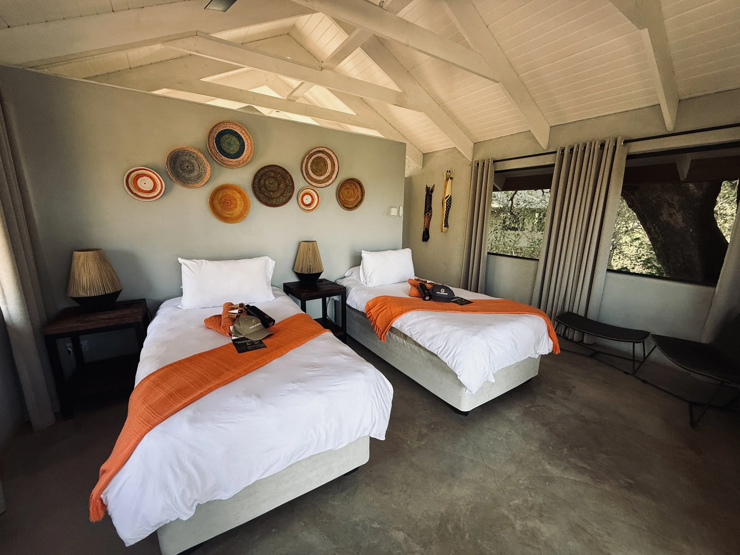 A cozy bedroom with two twin beds dressed in white bedding and orange throws. Each bed has a dark wood nightstand with a conical woven lampshade. The wall behind the beds displays colorful round woven art. There are two giraffe sculptures on the wall and large windows with beige curtains revealing lush greenery outside. The ceiling is white with exposed beams and skylights.