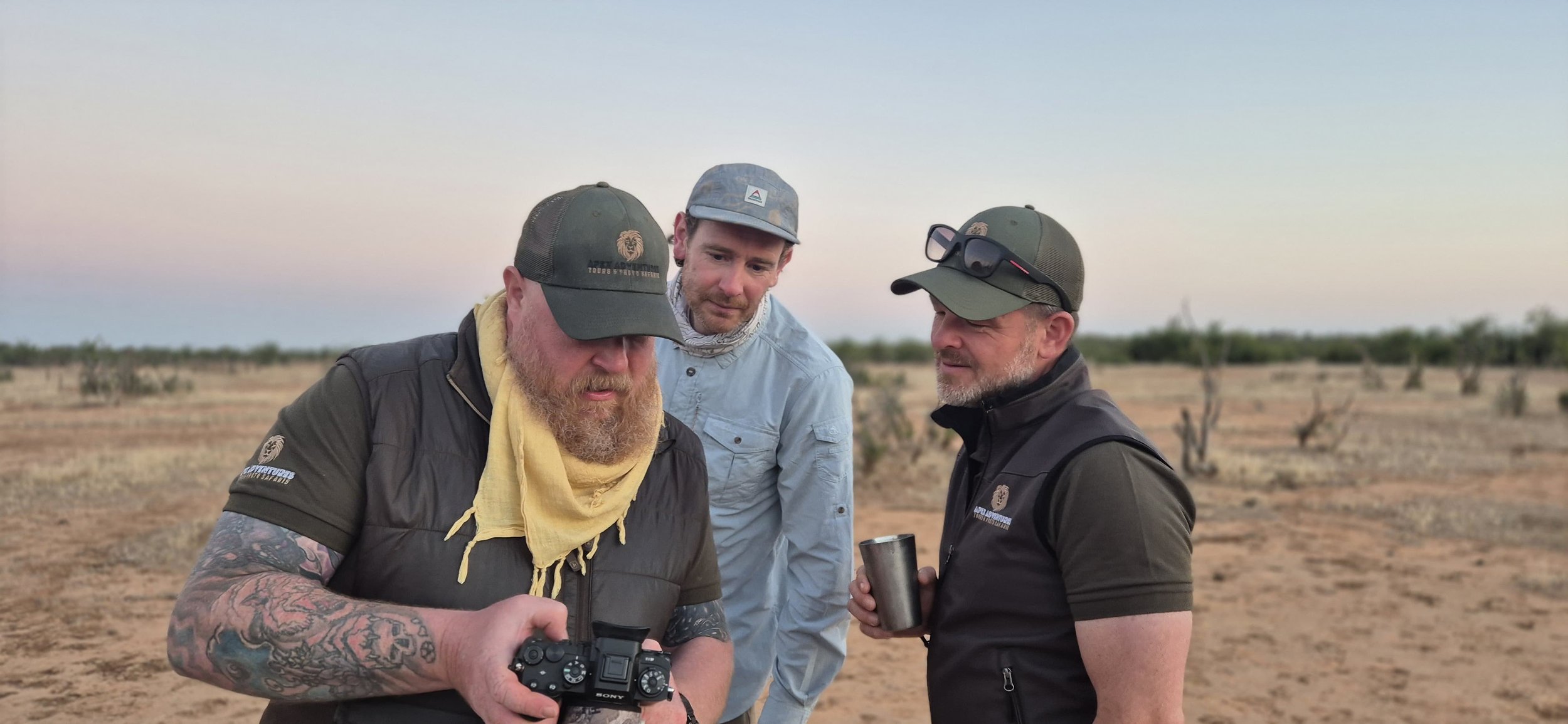 Three men in the desert looking at a camera, one holding a drink, all wearing outdoor gear and hats.