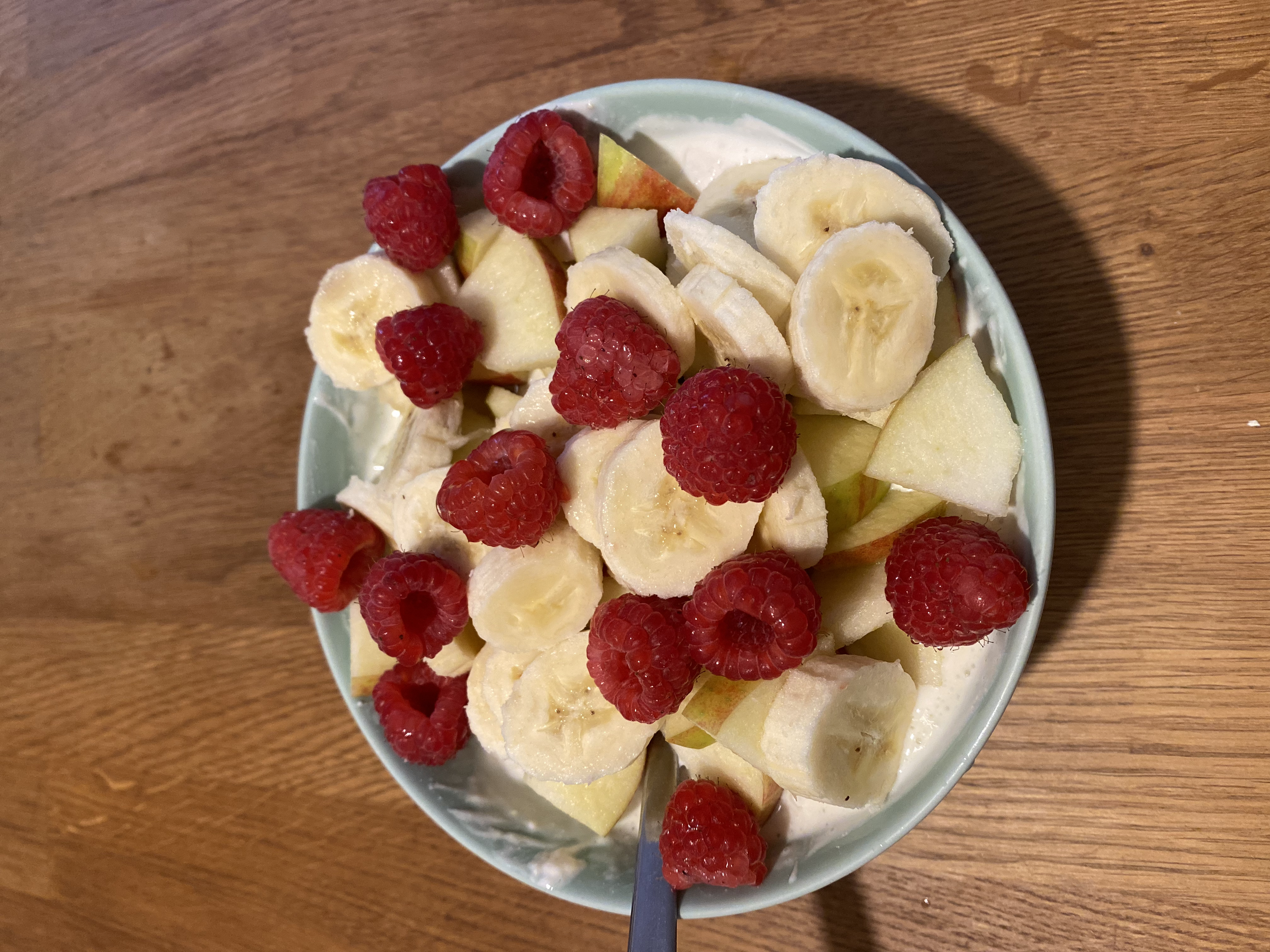 Schale mit geschnittener Banane, Himbeeren und Apfelstücken auf einem Holztisch.