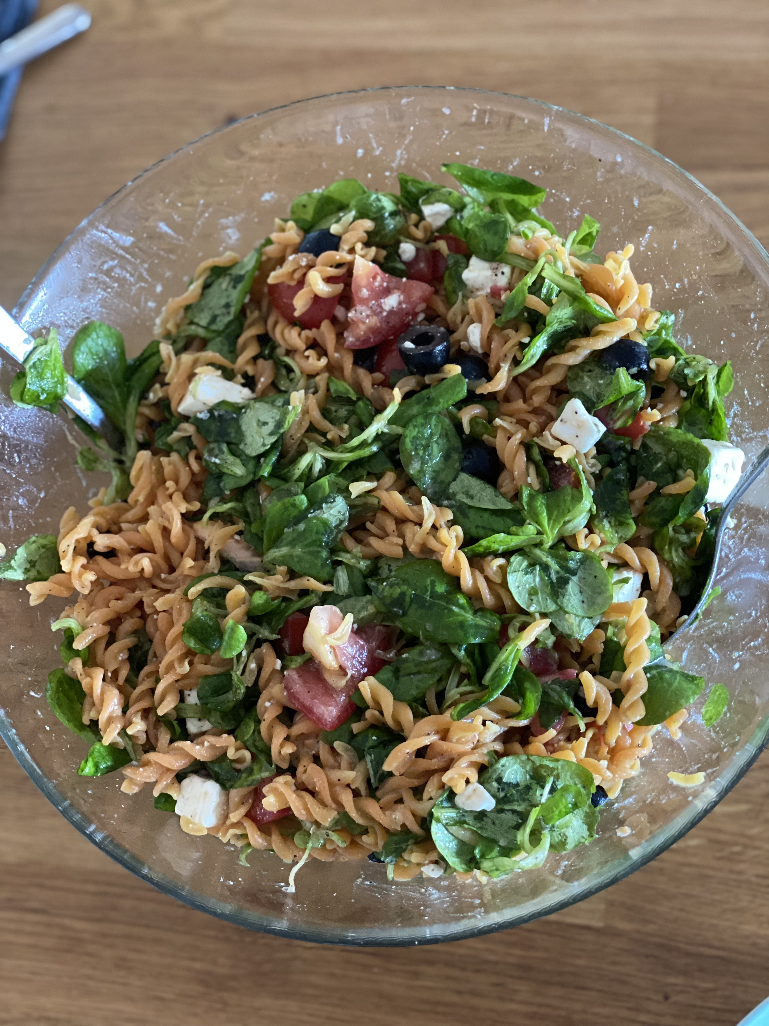 Schüssel mit Nudelsalat, der aus Fusilli, frischem Blattspinat, Tomaten, Oliven und Feta besteht, auf einem Holztisch.