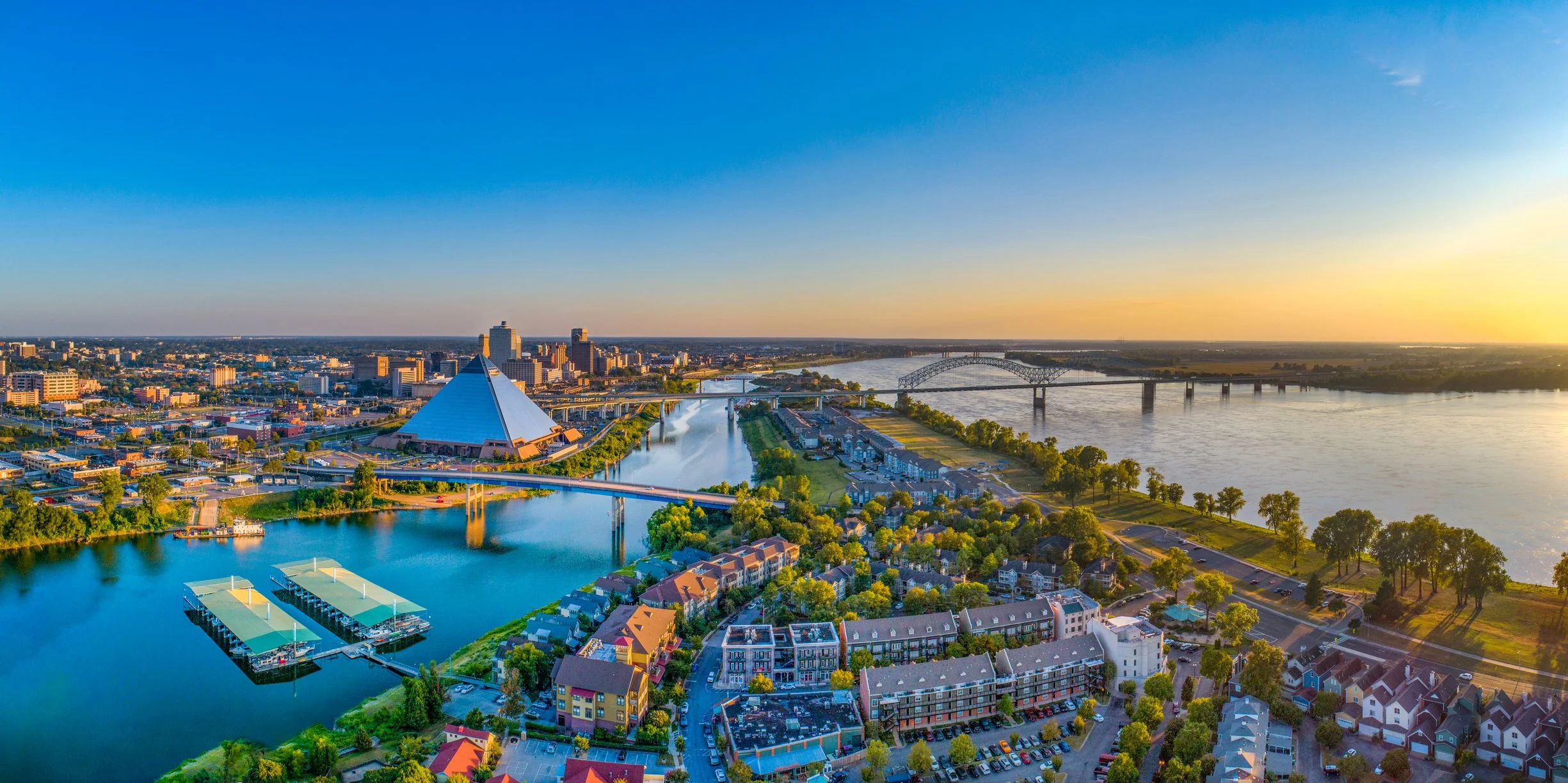 Aerial view of Memphis, Tennessee at sunset, showcasing the Mississippi River, a bridge, and the Memphis Pyramid stadium, with residential and commercial buildings.