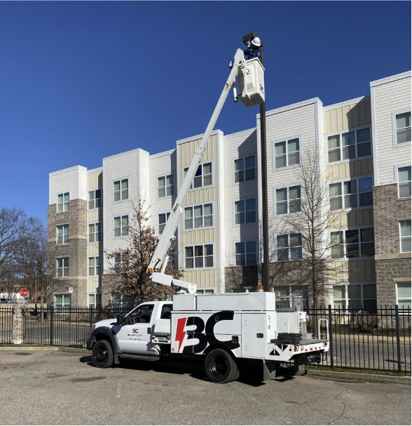 A utility bucket truck lifting a worker in a bucket to perform maintenance on a streetlight in front of a fenced residential building.