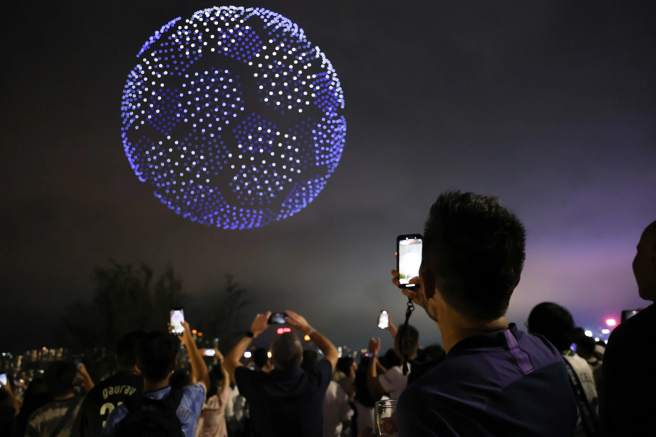 People watching a drone light show resembling a globe in the night sky, with many capturing photos on their phones.
