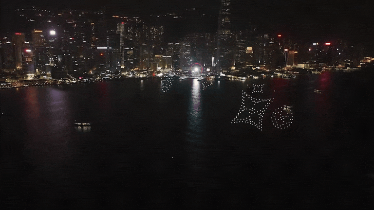 Nighttime city skyline with illuminated buildings across a river, and a large light display shaped like a star and the number 23 in the sky.