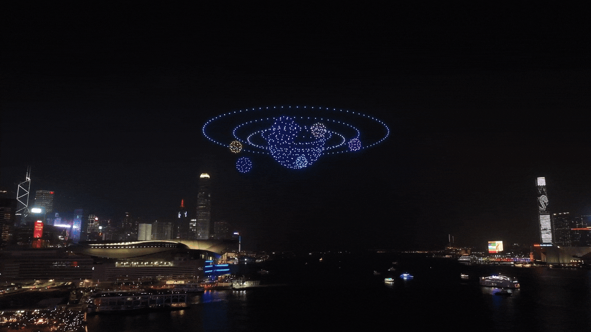 A nighttime city skyline with illuminated skyscrapers and a floating installation resembling a planet with rings and smaller planets orbiting, made of blue, purple, and orange lights.