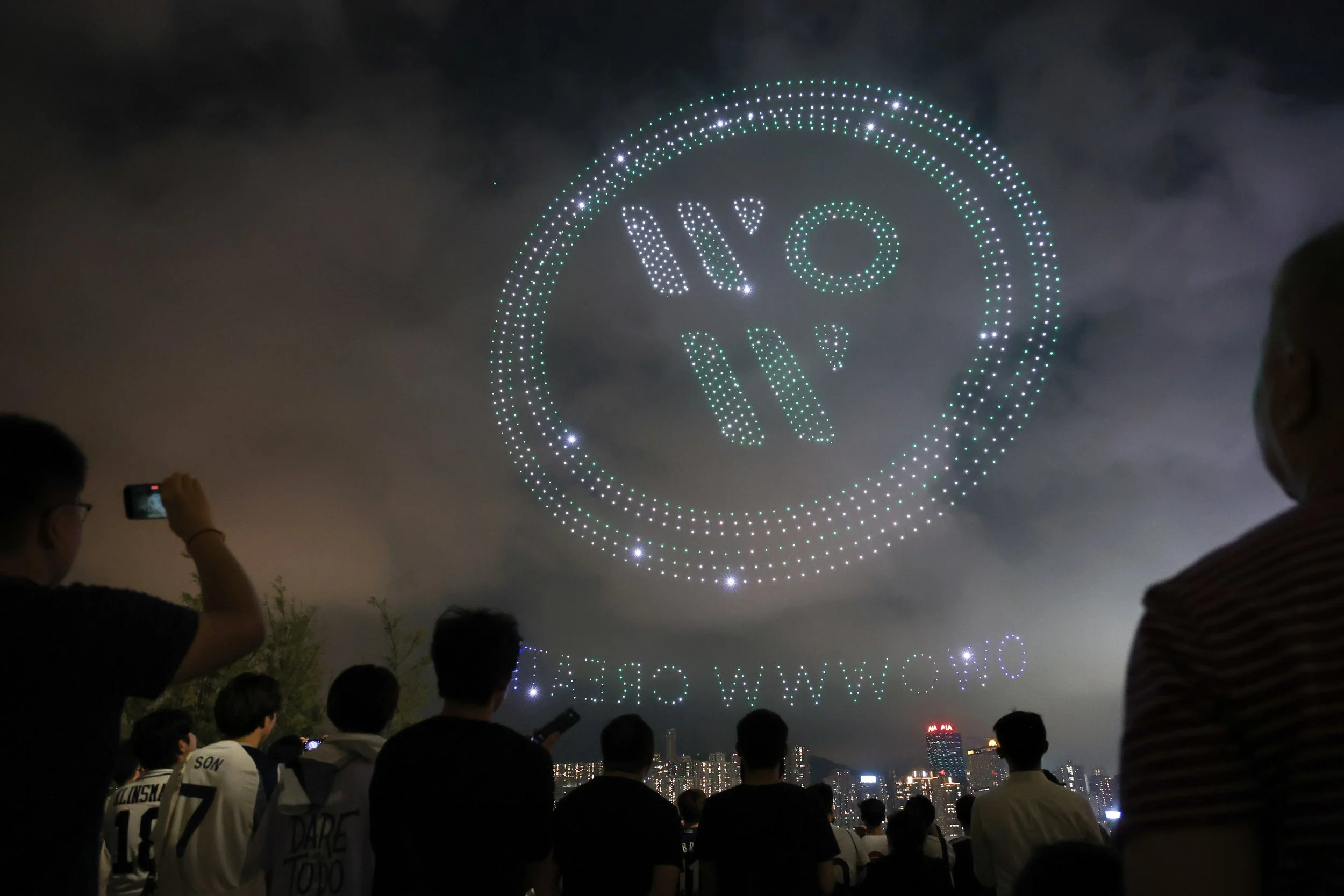 A drone light show illuminates the night sky with a large circular formation displaying a smiley face, hearts, and stylized text. People are watching, some taking photos, with a city skyline visible in the background.