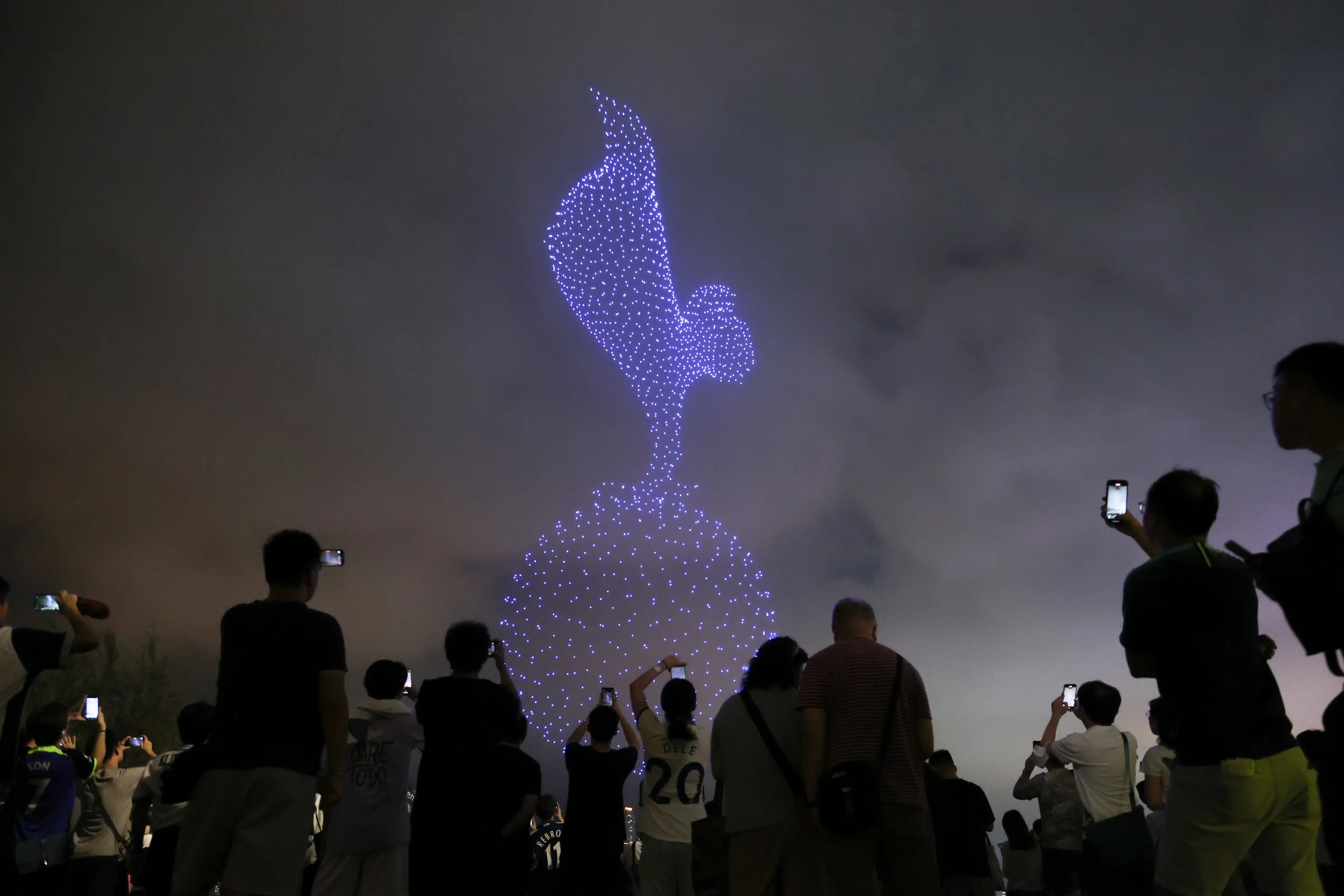 People gathered at night, taking photos of a large illuminated sculpture of a dove made of blue lights, with the background of cloudy sky.