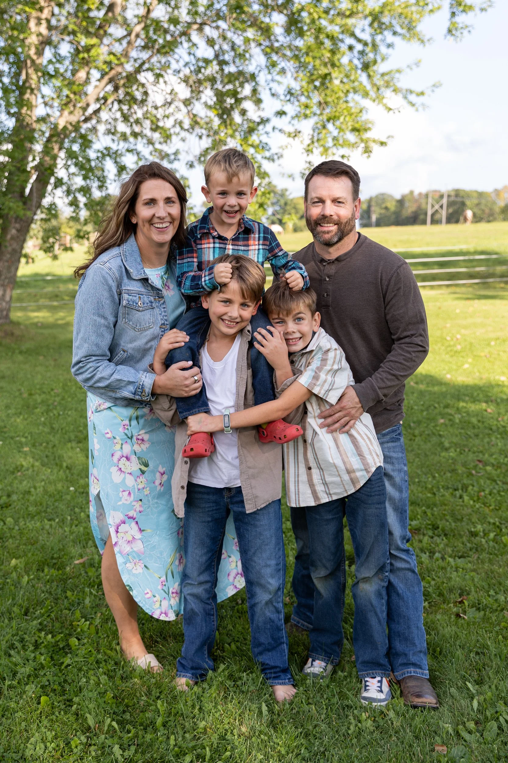 Family of five outdoors on a sunny day, standing on green grass with trees in the background, smiling at the camera.