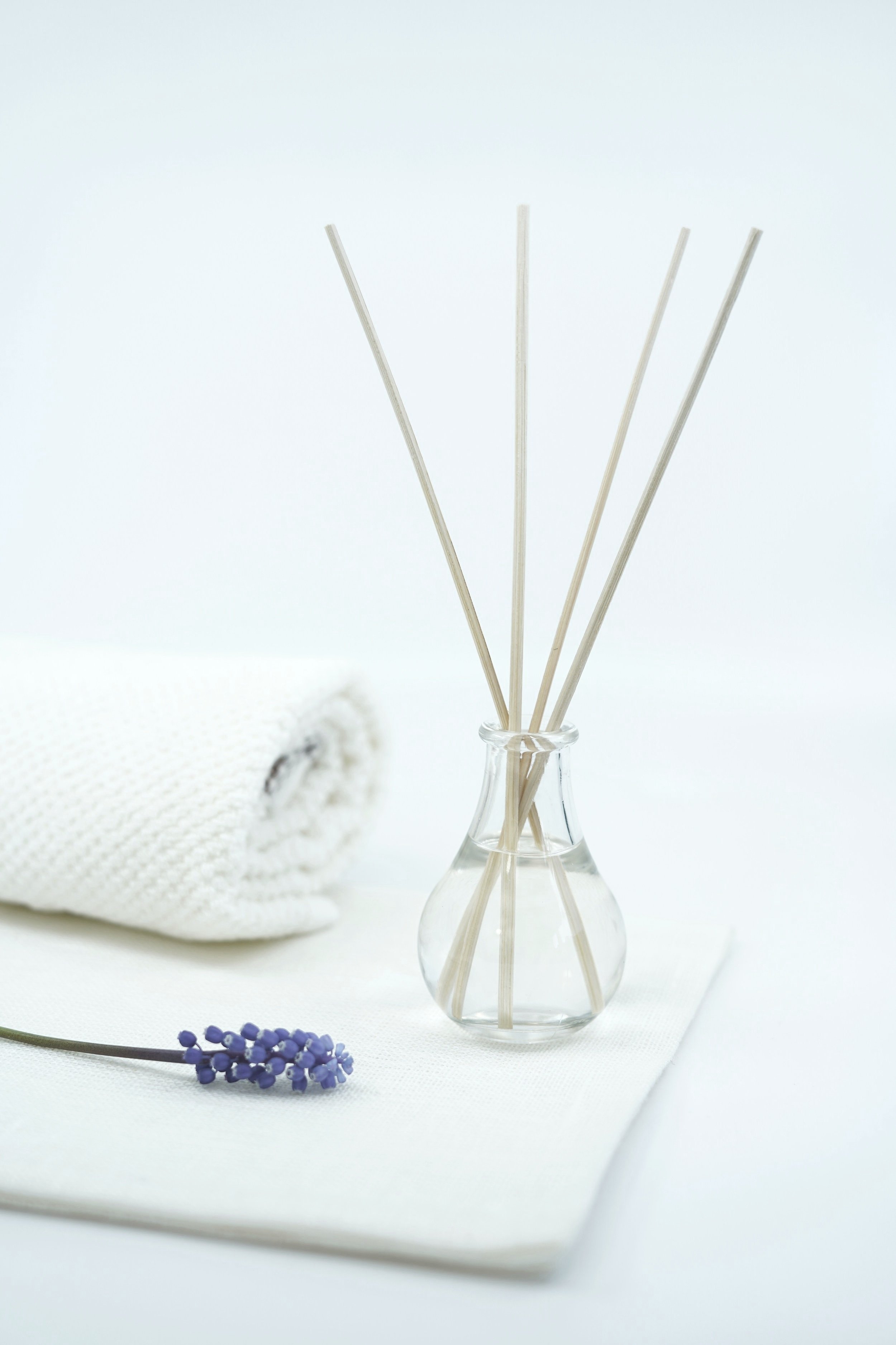 Aromatic reed diffuser in a glass bottle with wooden sticks, placed on a white towel next to a lavender flower.