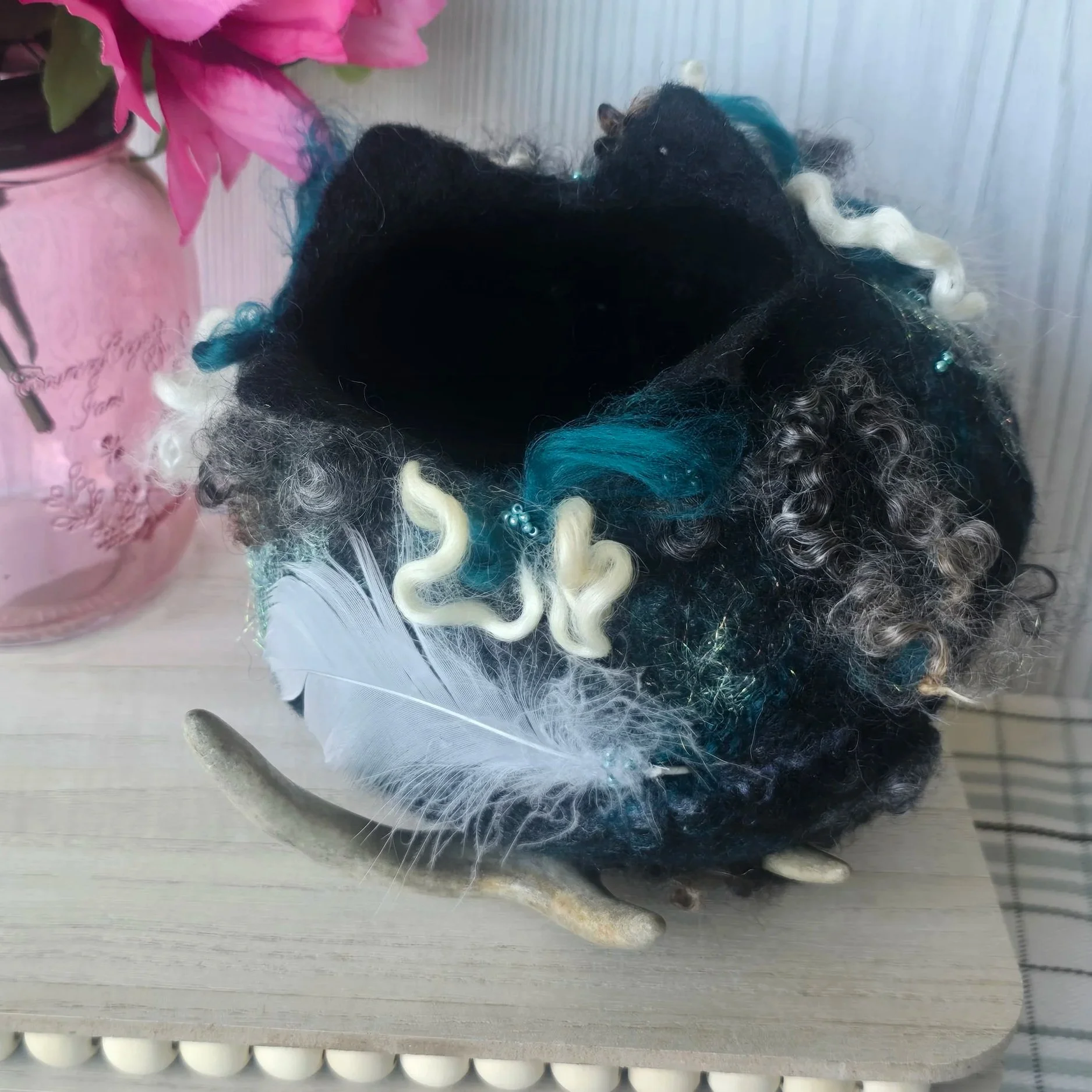 Colorful curly-haired wig with blue, white, black, and blonde curls, placed on a white surface with decorative feathers and beads, next to a pink decorative item.