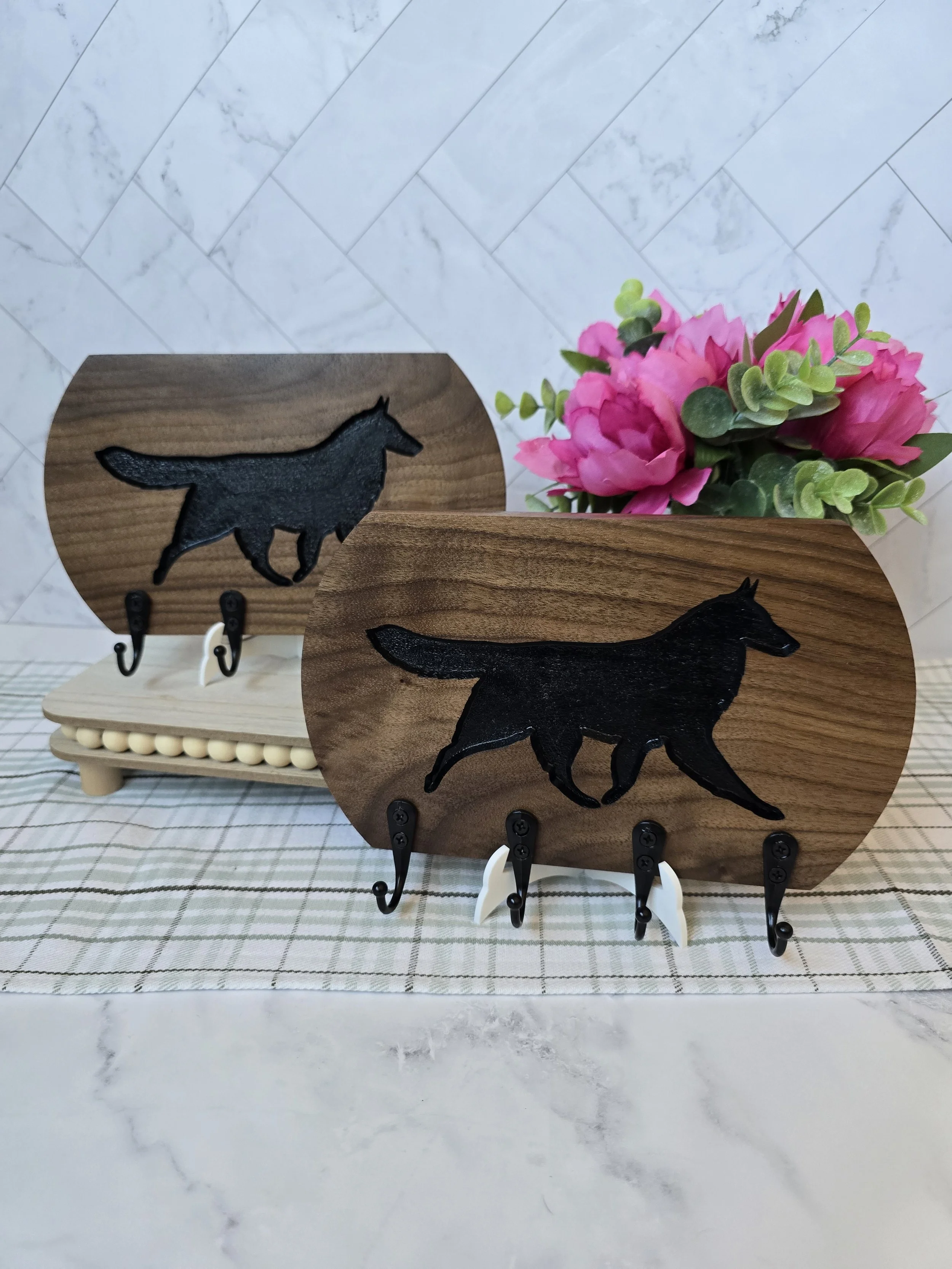 Decorative wooden plaques with black wolf silhouettes, hooks underneath, surrounded by pink flowers and green foliage, displayed on a checkered tablecloth.