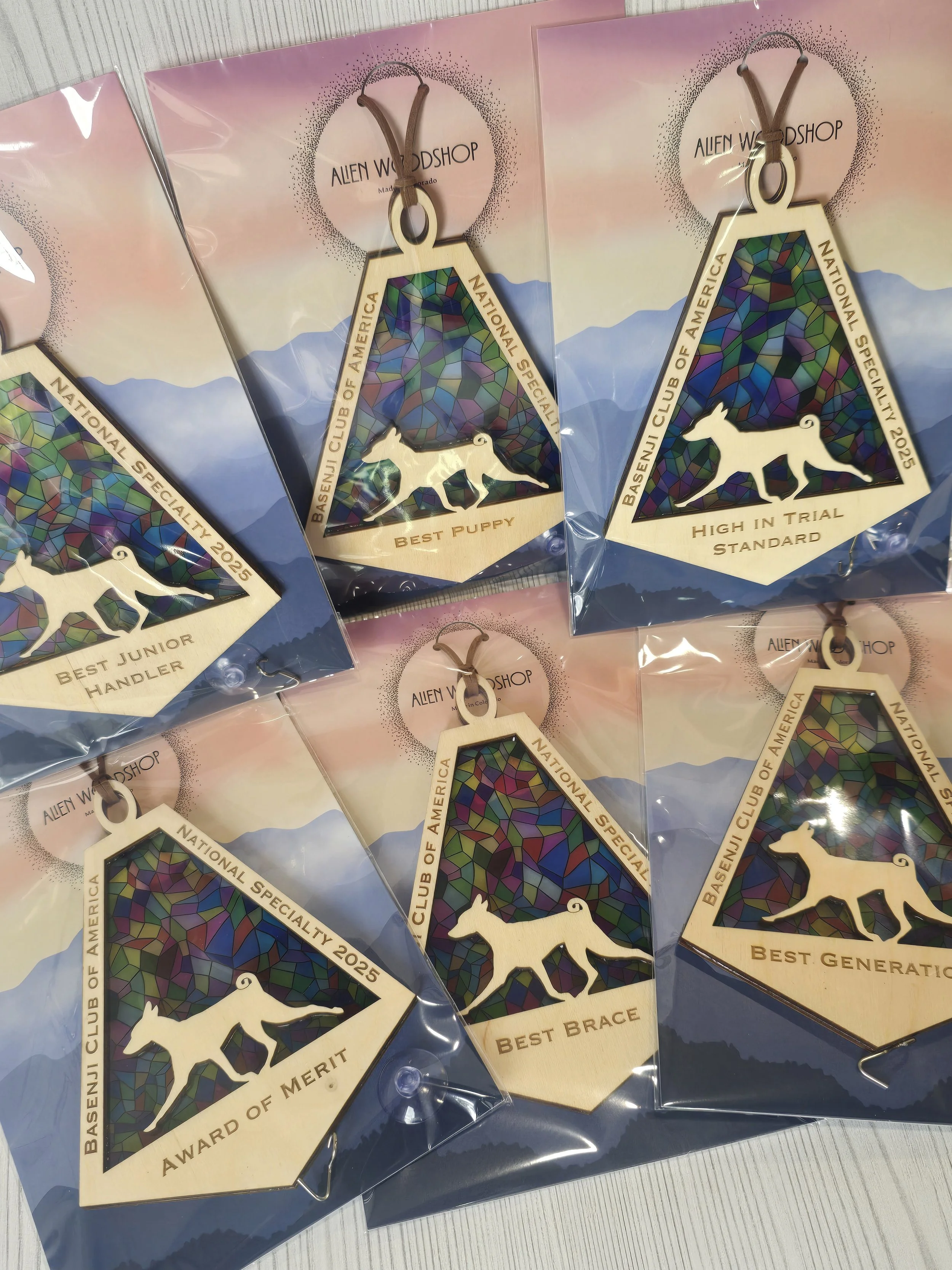 Recognition of several wooden keychains in plastic packaging featuring a mountain design made of colorful stained glass-like patterns, with a white silhouette of a dog, and inscriptions such as "Best Puppy," "High in Trial Standard," "Best Junior Handler," "Award of Merit," "Best Brace," on a silver surface background.