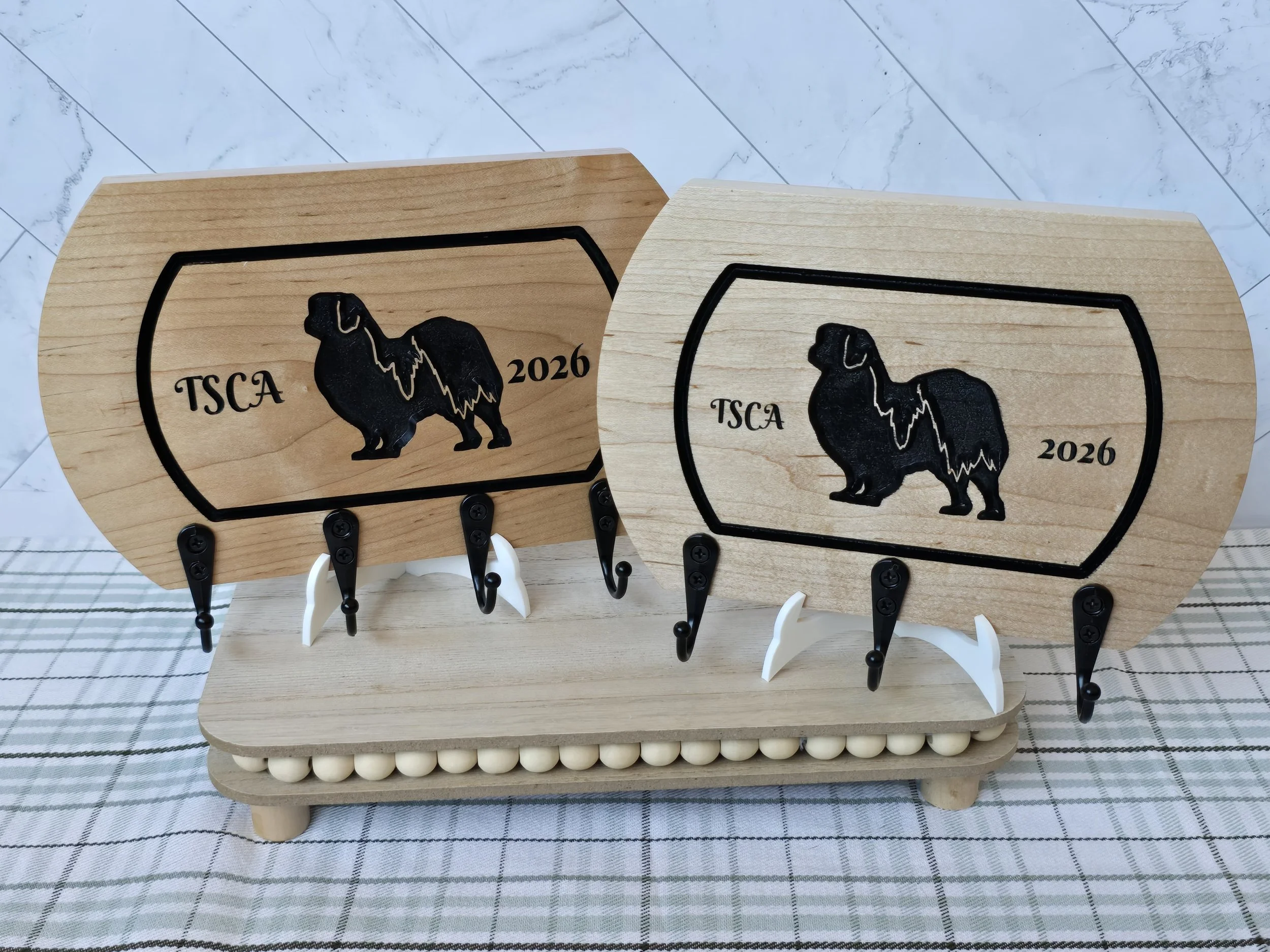 Two wooden leash holders with Tibetan Spaniel silhouettes and 'TSCA 2026' inscribed, displayed on a wooden stand with a checkered cloth and a marble floor background.