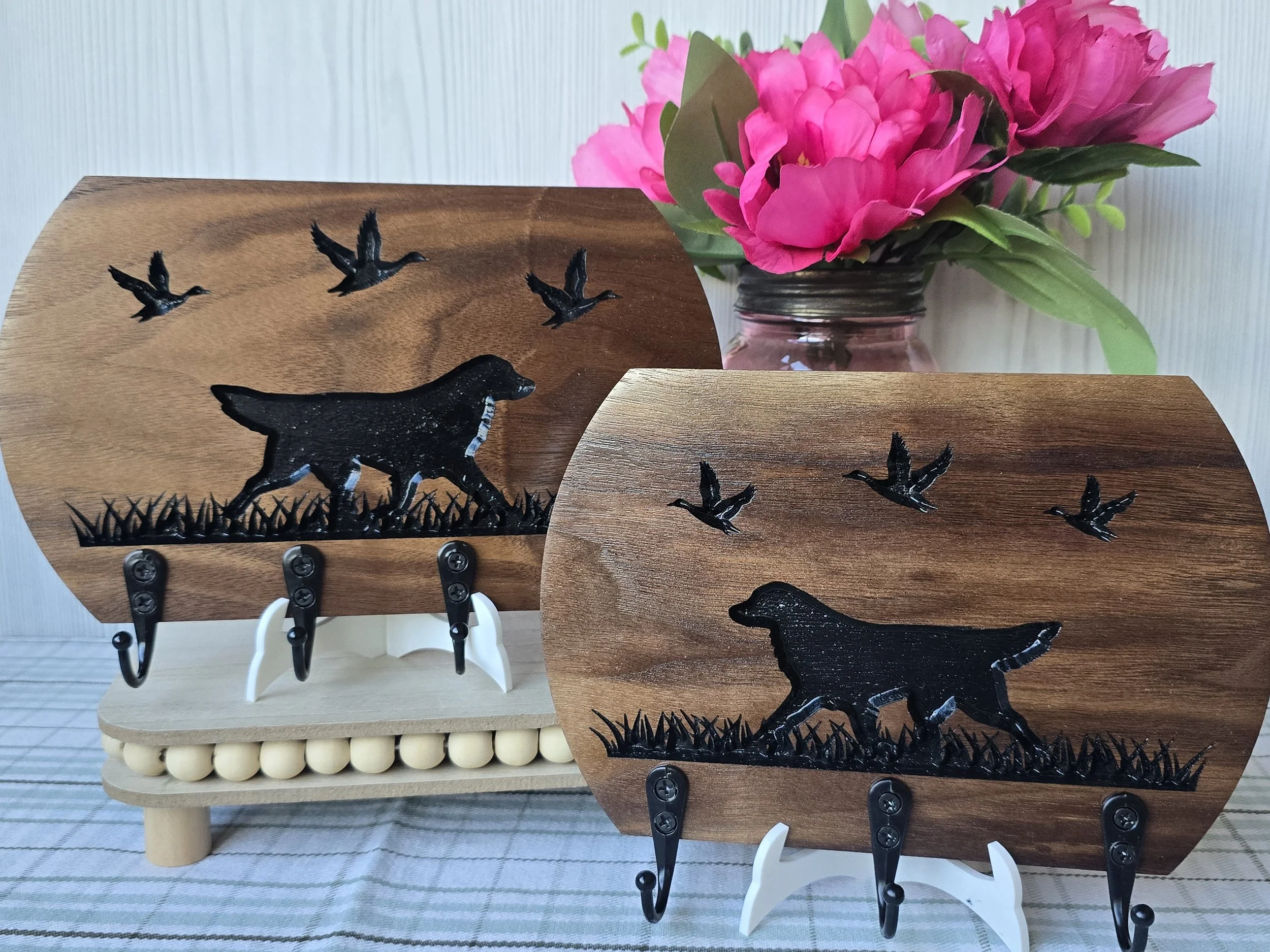 Decorative leash holder with Golden Retriever silhouettes with black hooks on a wooden stand with a background of pink flowers in a glass jar.